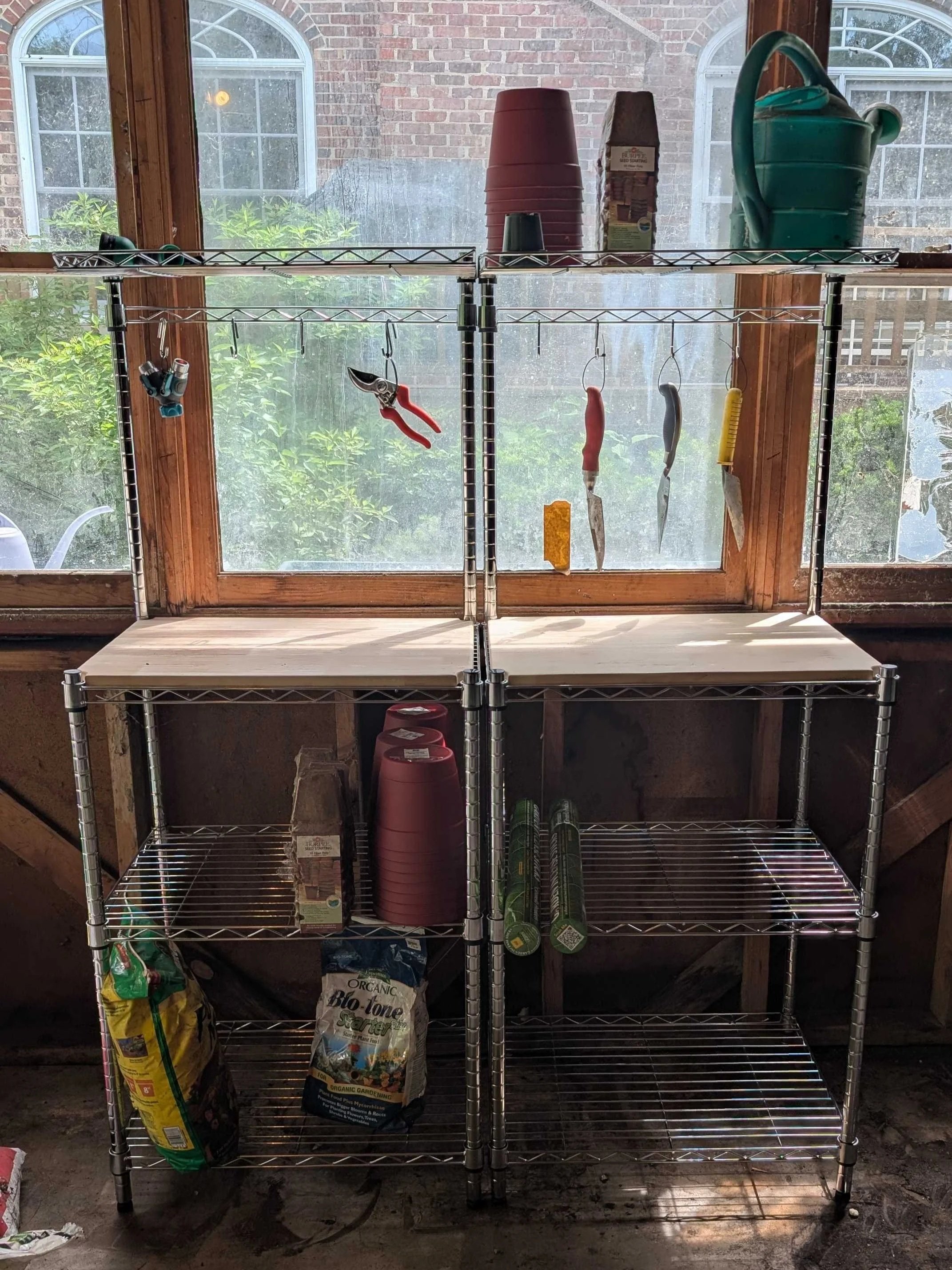Two shelving units with table top and gardening supplies stored on the shelving including dirt, shovels, and a watering can