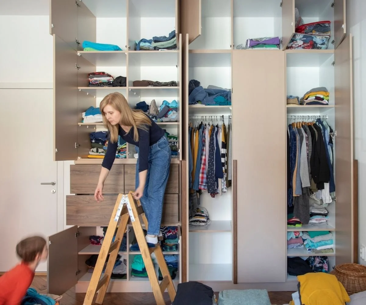 Image of a built in closet with all doors open showing folded and hanging clothing. A woman is standing on a small ladder and is bending down to grab something from a child sitting on the floor.