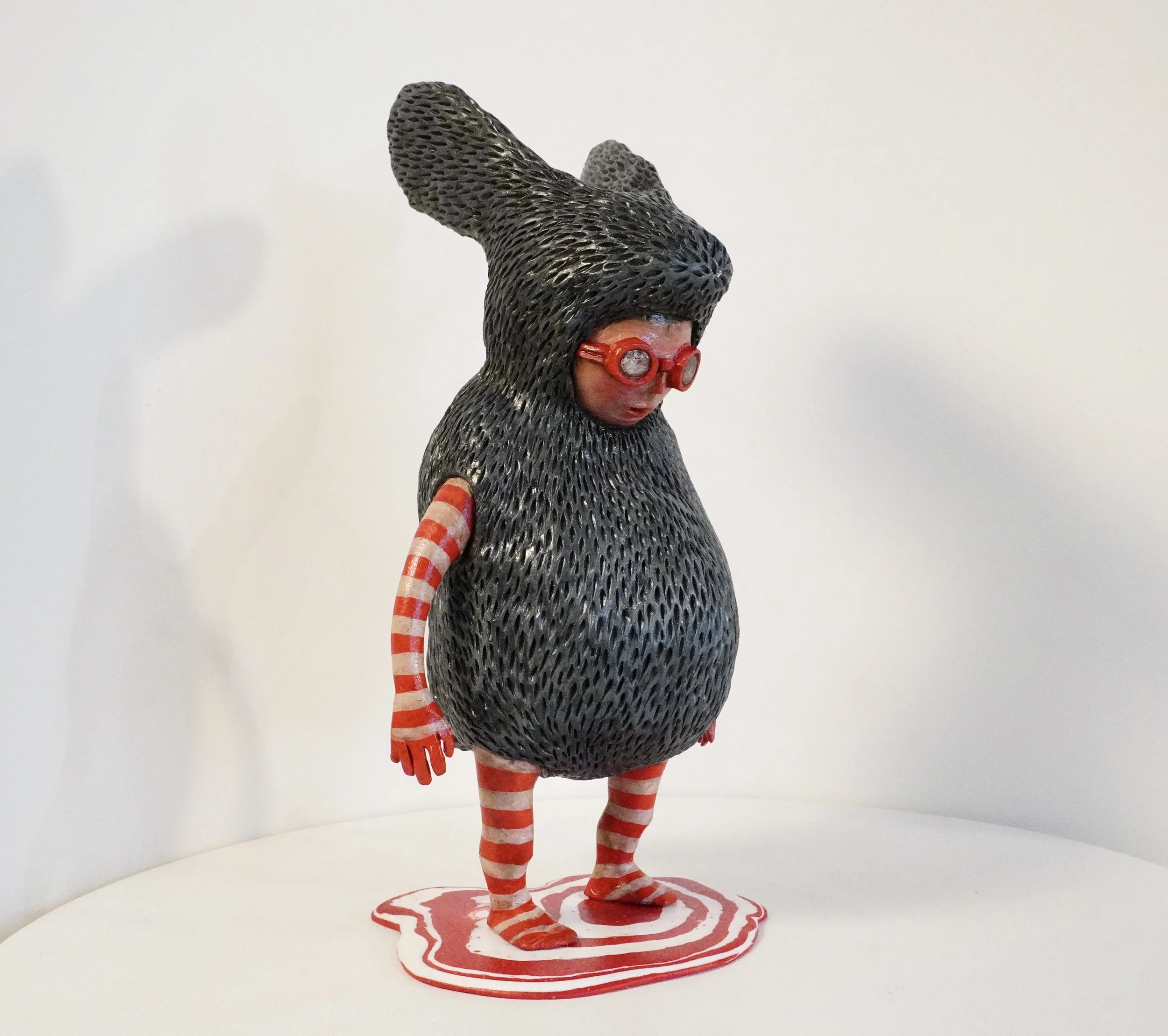 Art sculpture of a girl with a large, textured, dark gray hat and glasses. The girl has red and white striped arms and legs and is standing on a circular red and white swirl base.