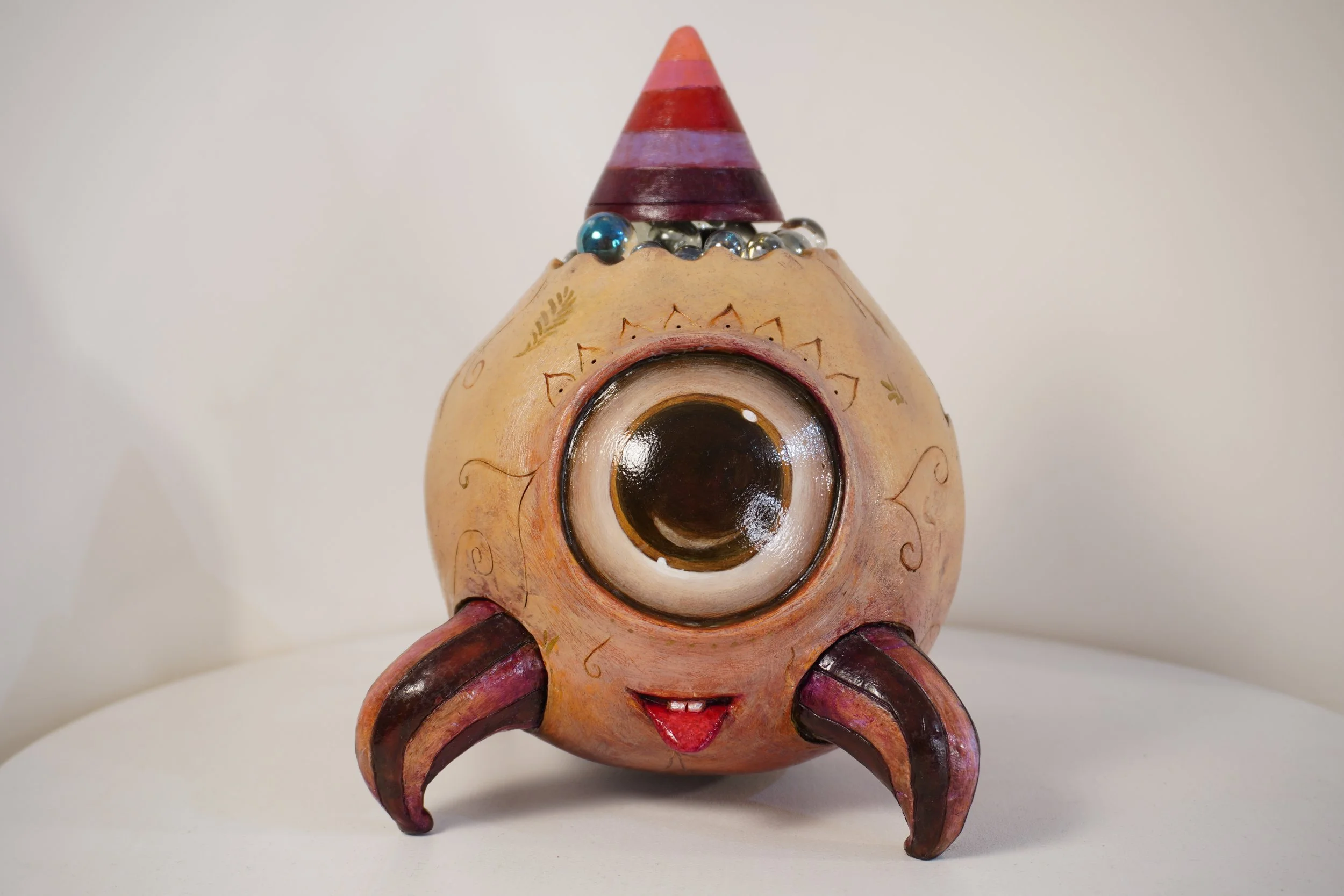 A decorative ceramic sculpture of a stylized character with a large eye at the center, a smiling mouth, and curved horns or ears, painted in earthy tones with purple and red accents.
