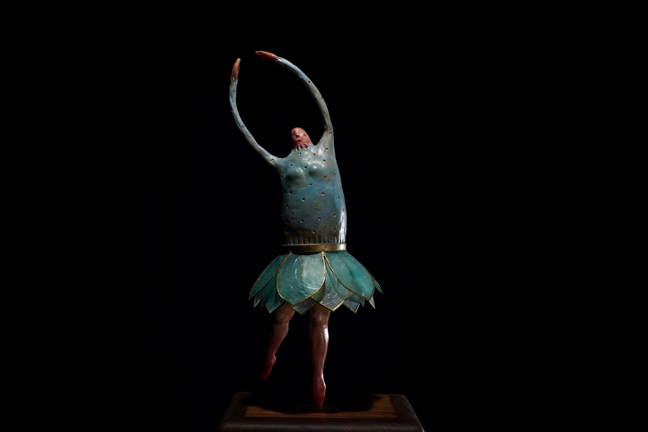 A sculpture of a woman with an elongated arm raised above her head, wearing a bluish-green dress with leaf-like details, set against a black background.