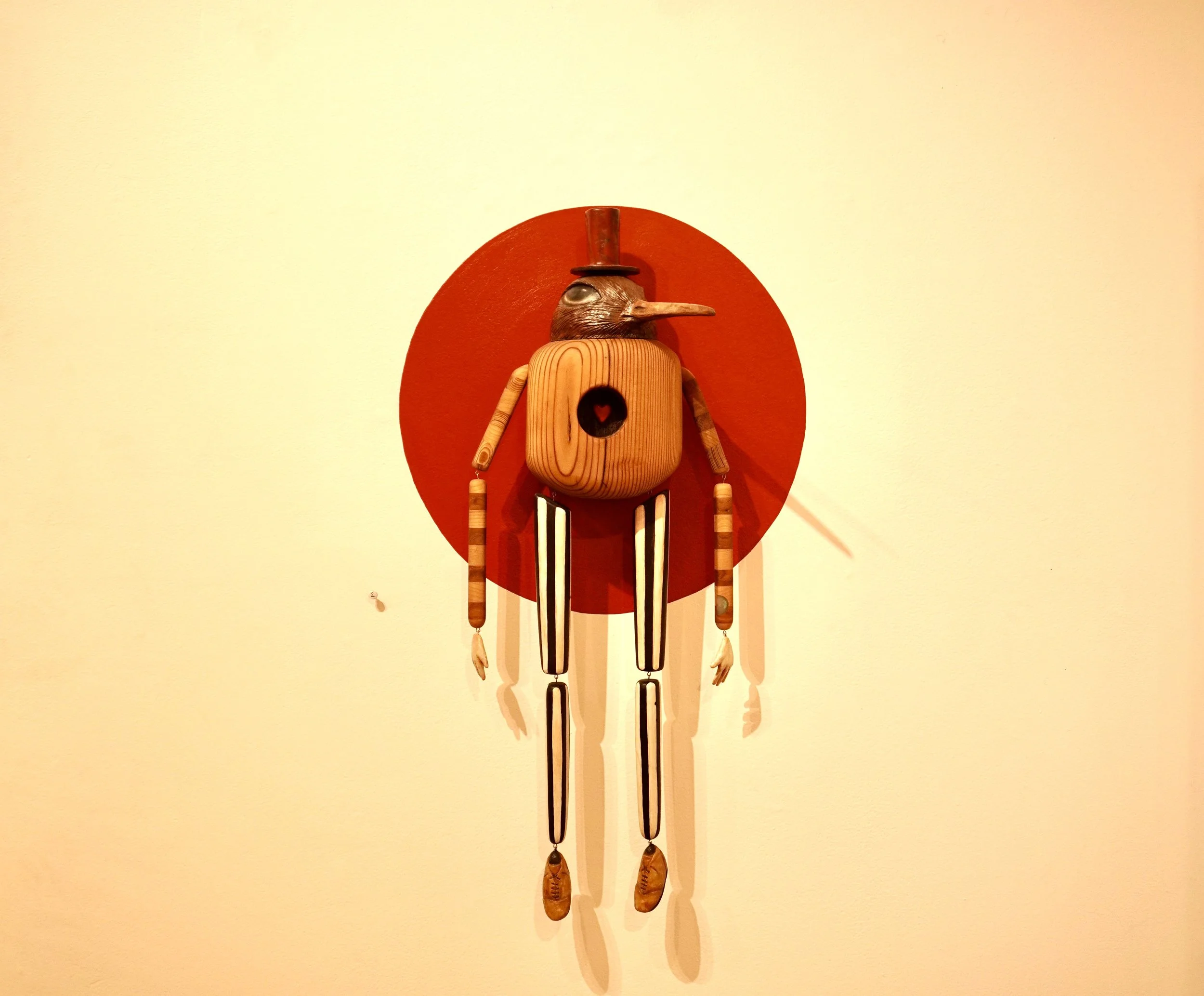 A whimsical wooden bird with a long beak and a small top hat, adorned with various wooden and metallic parts, standing against a red circular background on a light-colored wall.