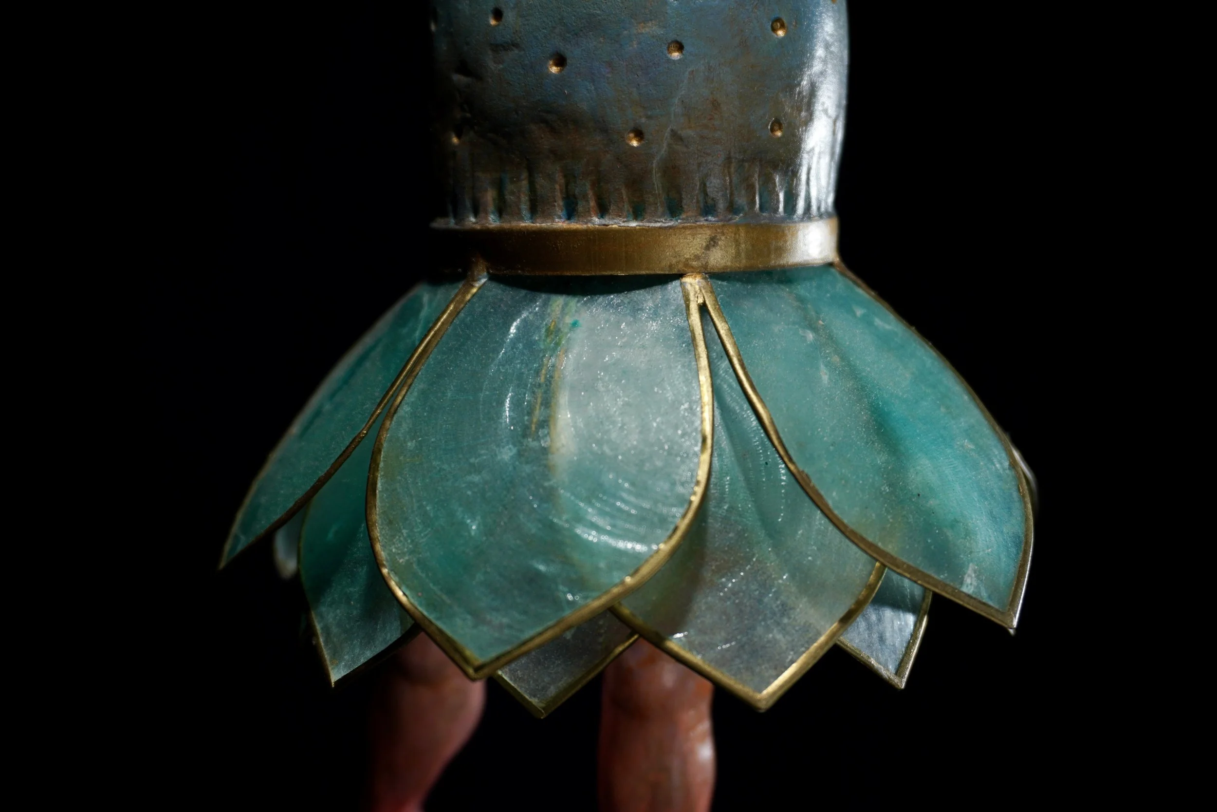 Close-up of a vintage green metal object with gold accents, featuring small holes and decorative gold wire, set against a black background.