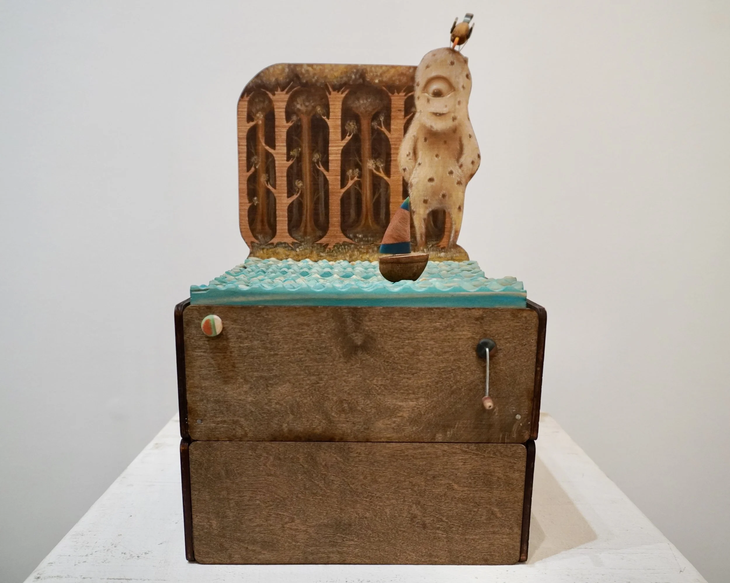 A kinetic piece made out of salvaged wood, featuring a roof with a crow decoy, a textured front wall with tree-like patterns, and a whimsical figure resembling a slice of bread with eyes and a mouth.