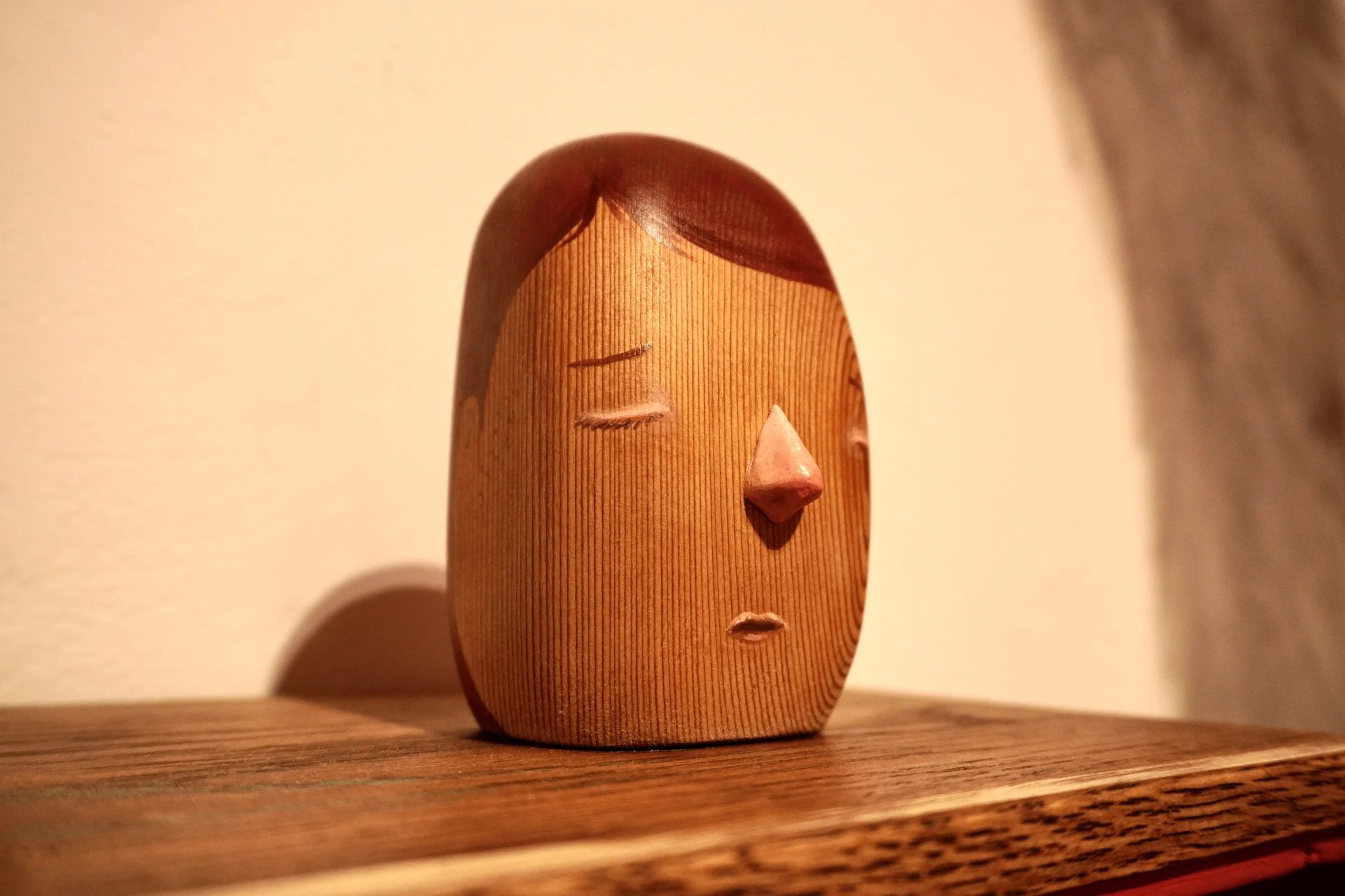 A carved wooden sculpture of a human face with closed eyes, a nose, and a serene expression, placed on a wooden surface.