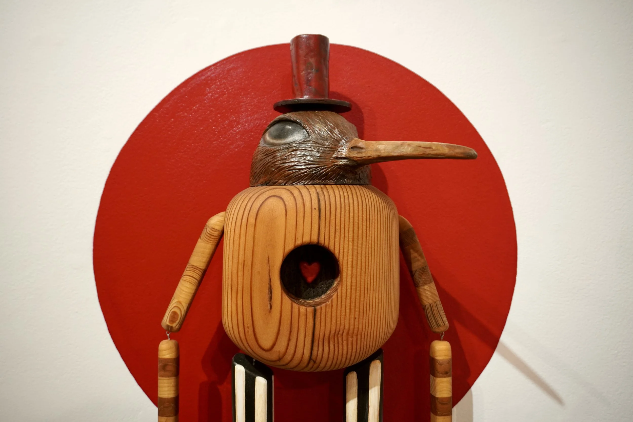 A wooden bird-shaped sculpture with a long beak and a top hat, mounted on a red circular background against a white wall.