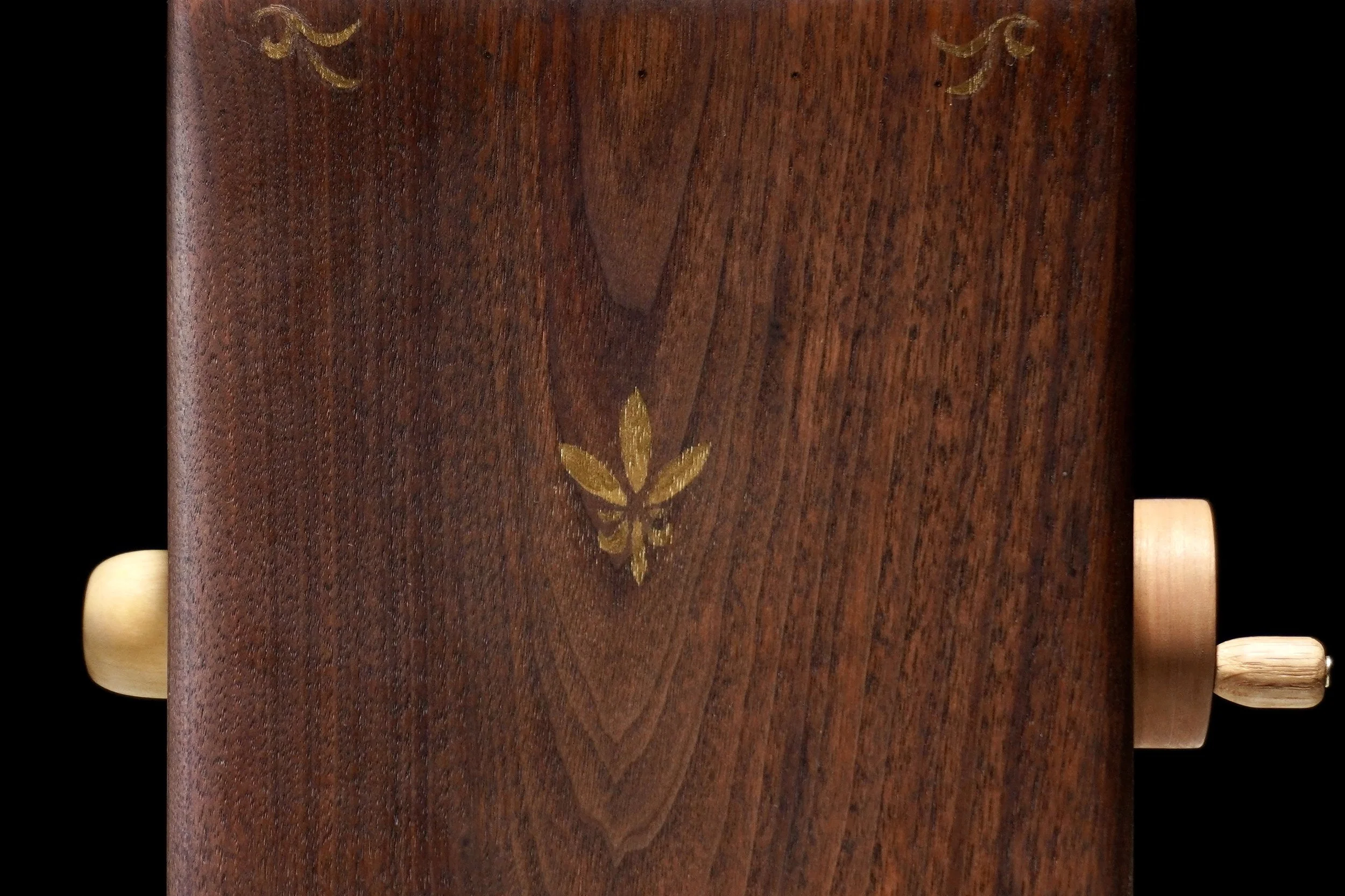A close-up of a wooden piece of furniture with a gold inlay of a flower or leaf design on its surface. The edges of the furniture are rounded, and there are small wooden pegs or knobs on the side.