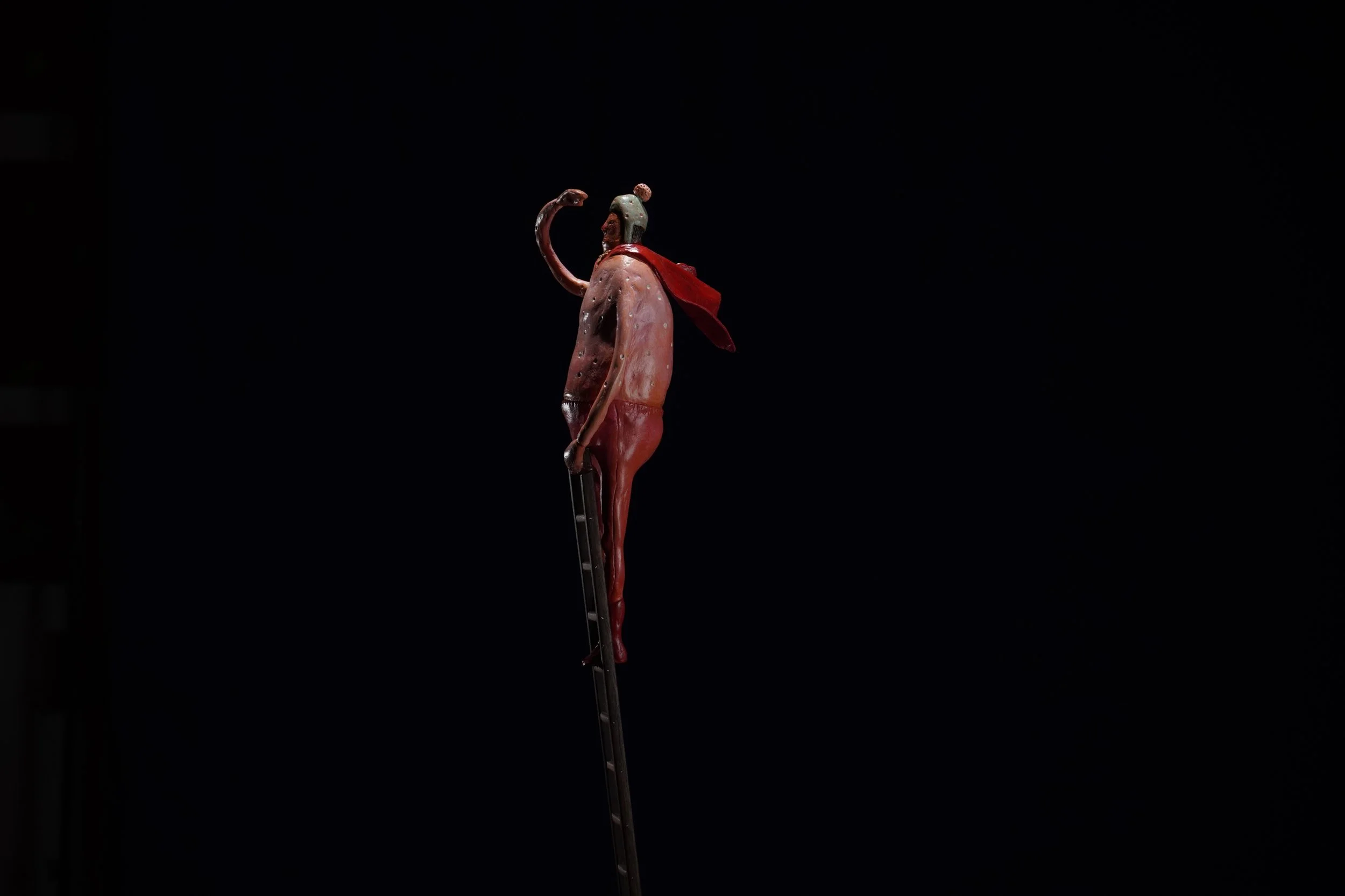 A sculpture of a human figure  wearing a red cape, standing on a thin ladder against a black background.