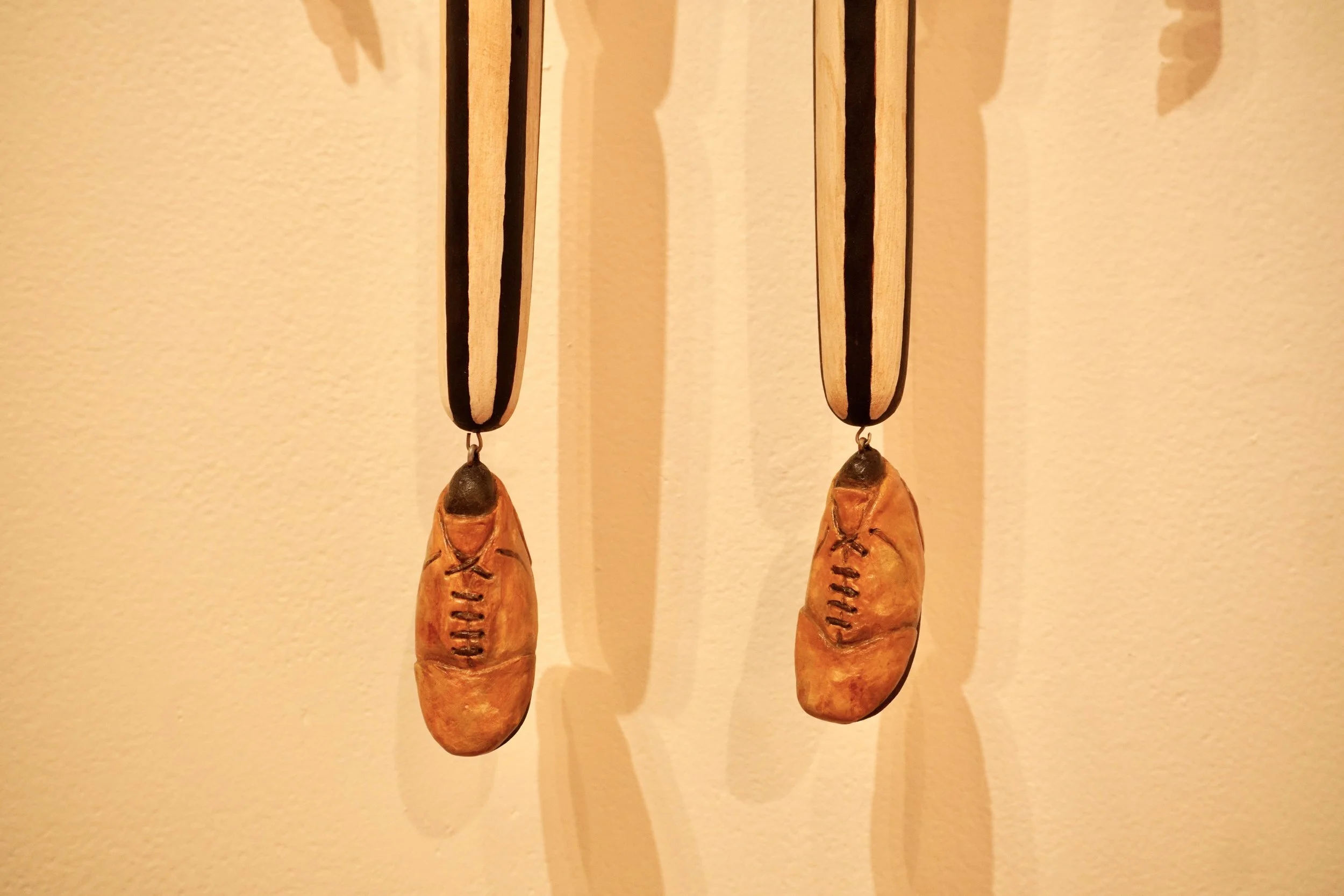 Close-up of two hanging decorative objects resembling football shoes, made of leather and wood, with their shadows cast on a beige wall.