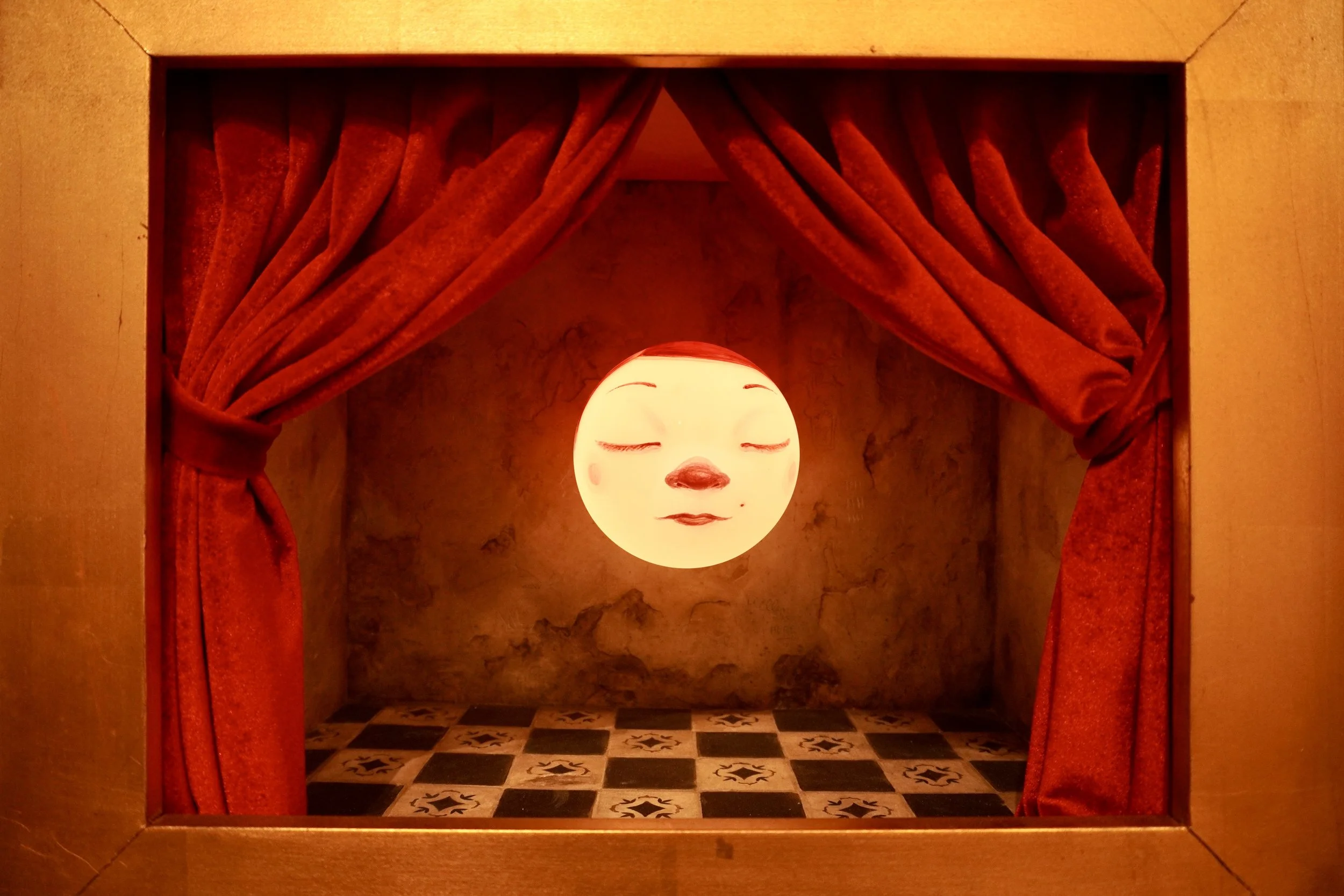 A glowing face mask with closed eyes and a slight smile, framed by red curtains in a small staged setting with a tiled floor.