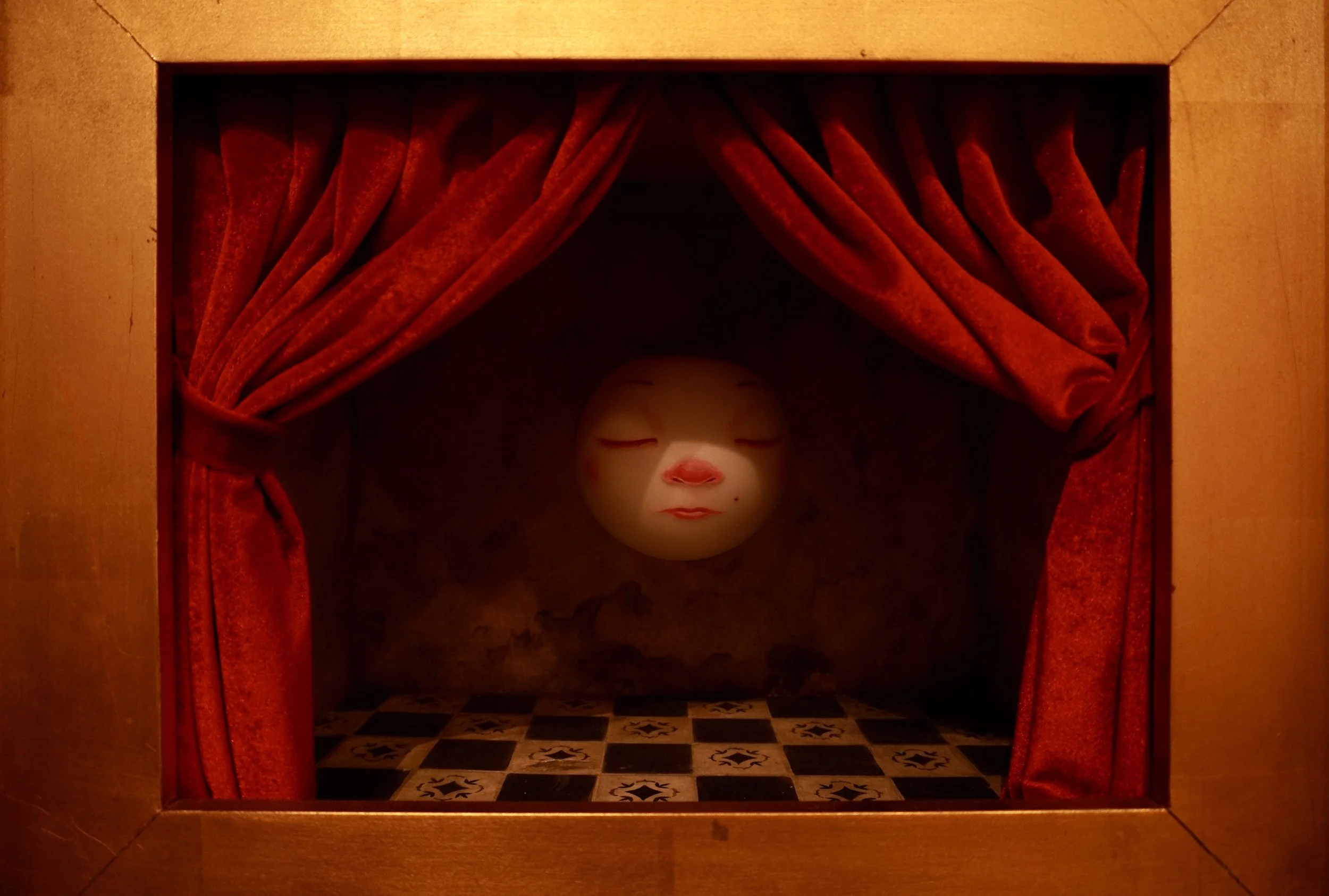 Small puppet with closed eyes and red lips in a shadowed, red velvet curtain setting.