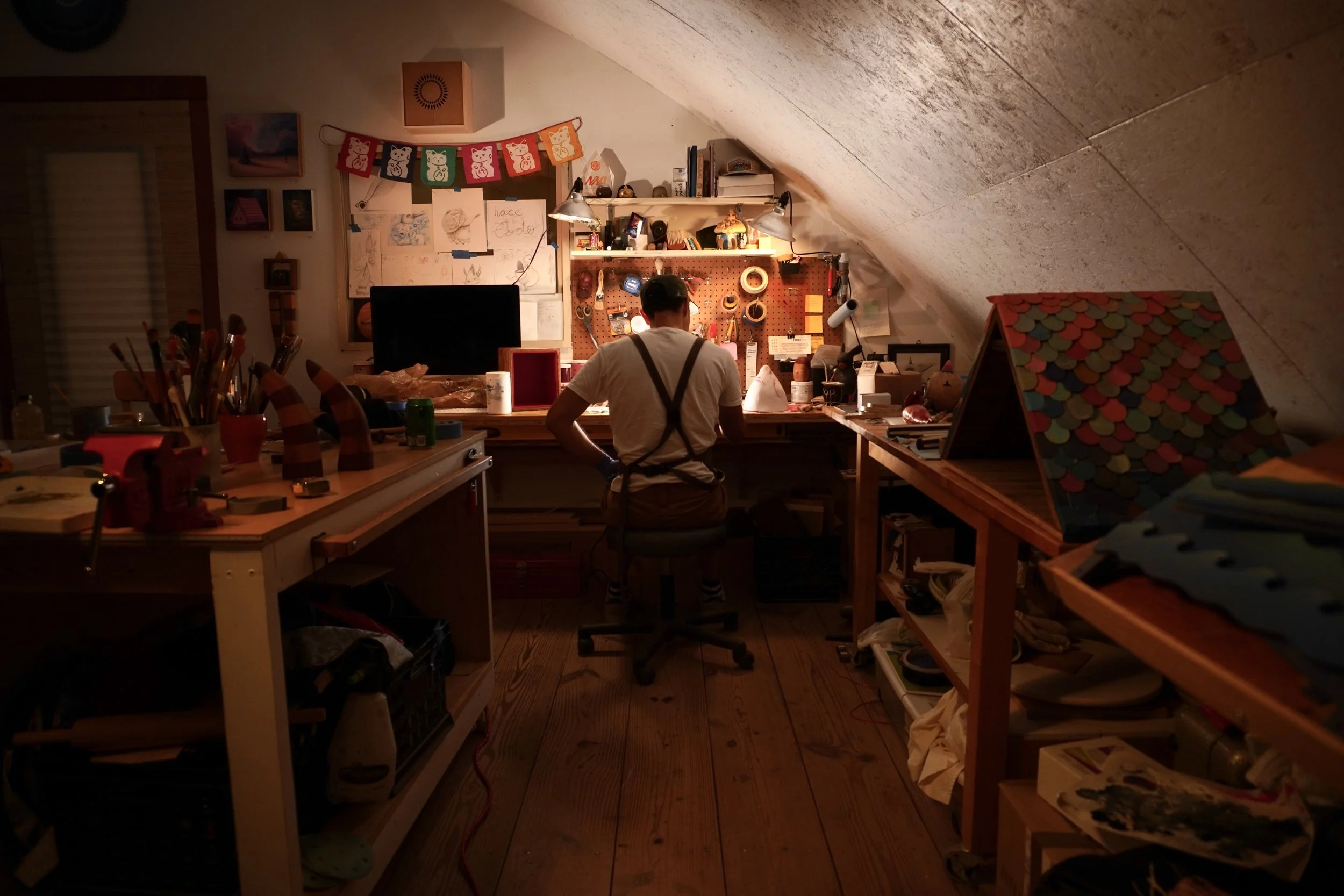 A person standing at a cluttered wooden desk in a cozy attic workspace with sloped ceiling, illuminated by desk lamps, with shelves of art supplies, books, and decorations behind.