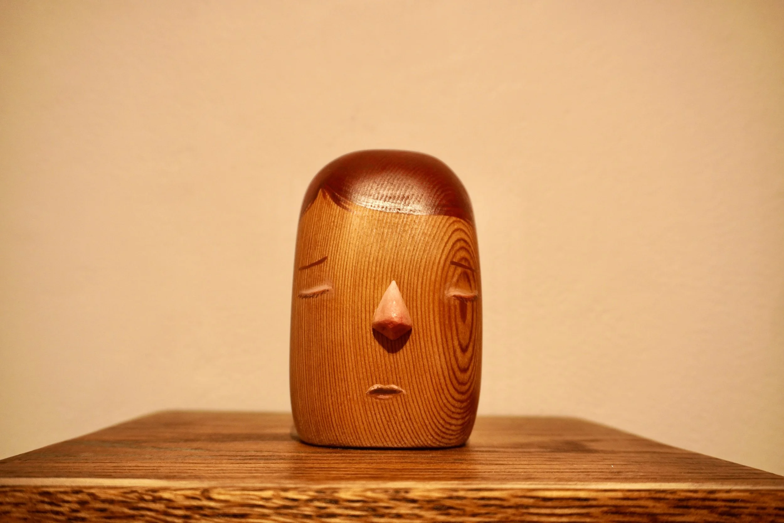 A wooden sculpture of a human face with closed eyes, a nose, and simplified lips, set against a plain background.