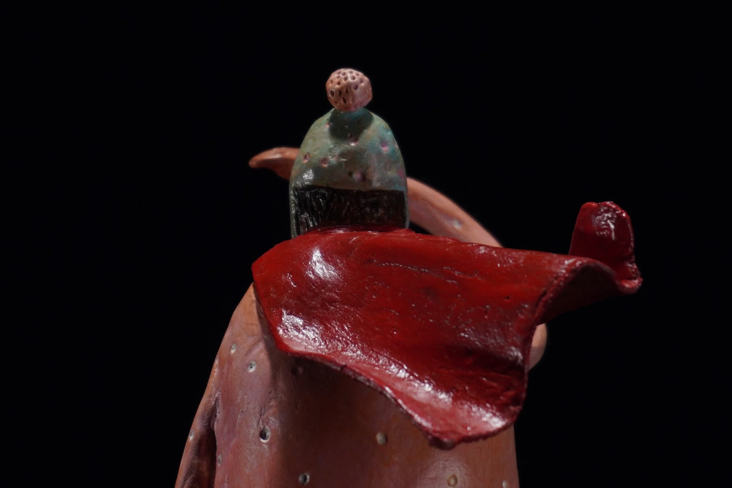 A close-up of a small decorative sculpture resembling a humanoid figure with a round head with holes, wearing a multicolored, shiny garment, seated on a red, irregularly shaped base, against a black background.