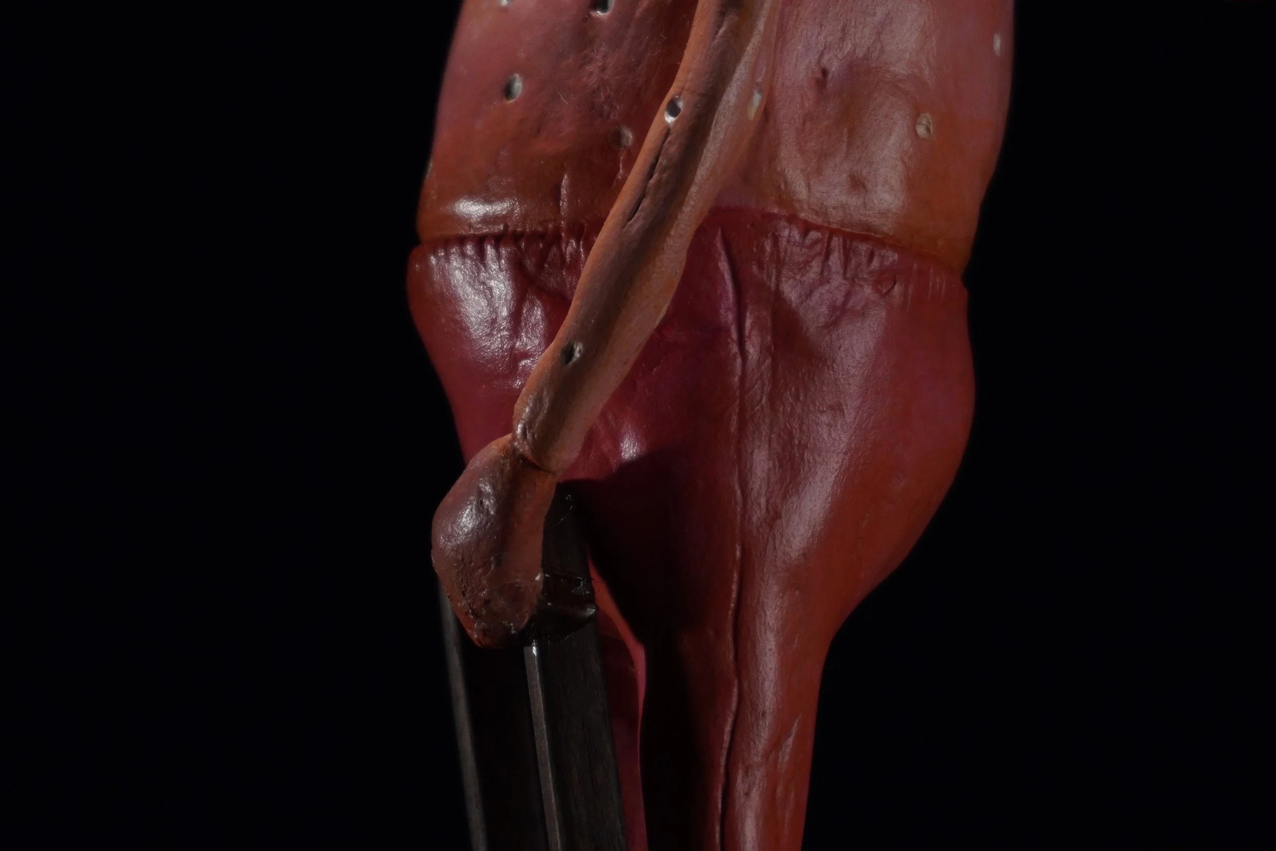 Close-up of a reddish-brown wooden mask or sculpture with a diagonal piece of wood running across it, set against a black background.