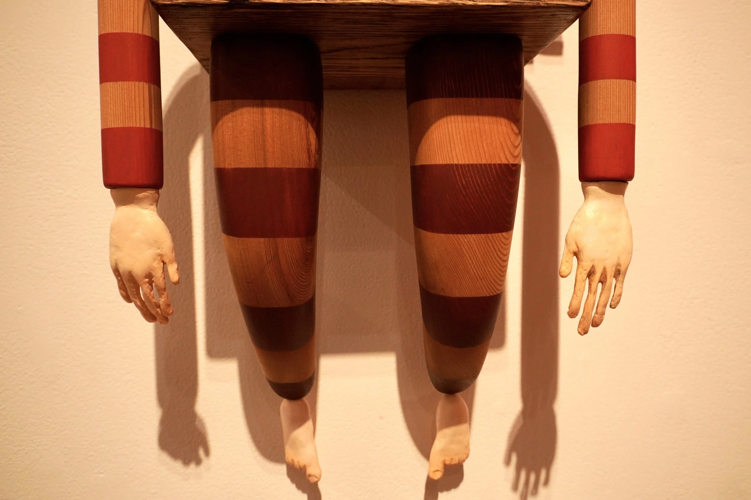 A mixed media artwork featuring a mannequin torso with arms, painted with horizontal red and natural wood-colored stripes, and a wood shelf mounted above it.