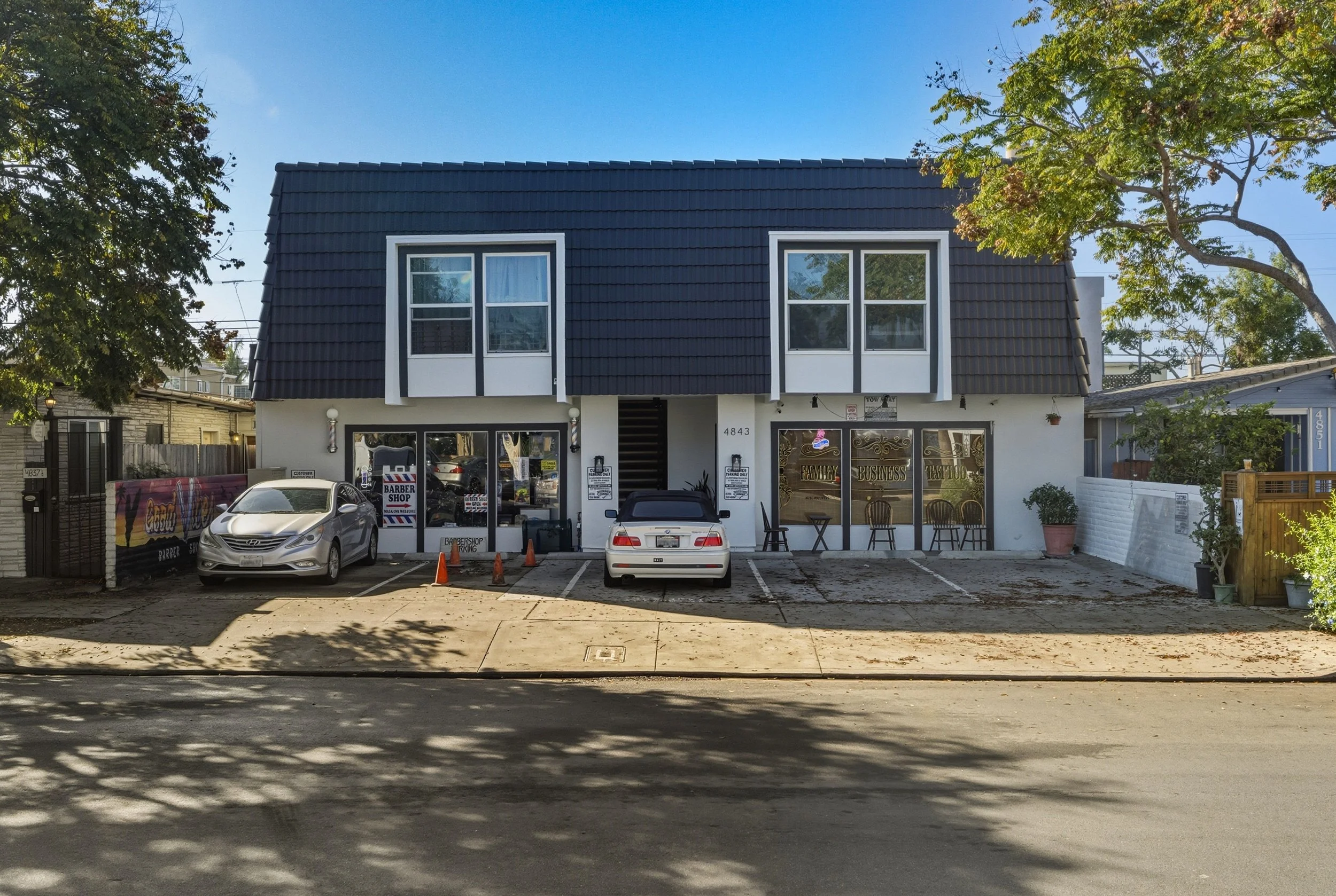 A two-story building with a blue tiled roof, white and black exterior, and a storefront on the ground level. There are two cars parked in front of the building, orange cones, and outdoor seating with chairs and tables. Trees with green leaves flank the building on the left and right sides.