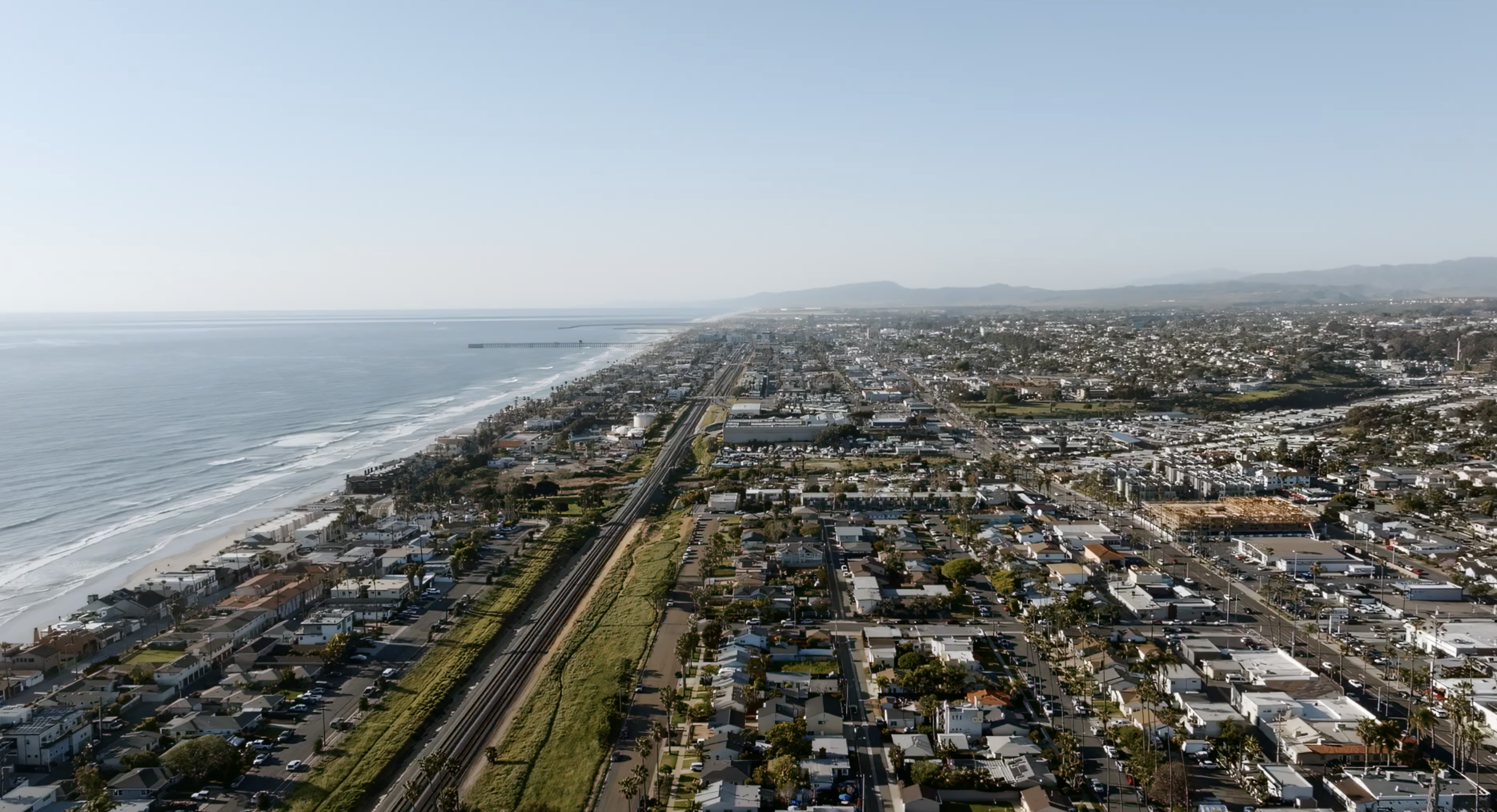 Aerial view of a Oceanside, California with residential and commercial buildings, a railway track, and a beach along the shoreline, with distant mountains under a clear blue sky.