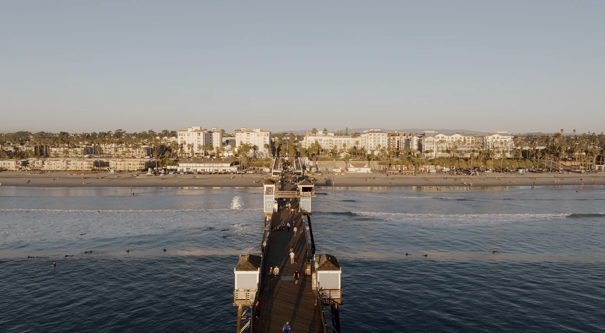 Oceanside pier extending into the ocean with people walking on it, leading to a beach and a row of buildings, including hotels, with palm trees in the background under a clear sky.