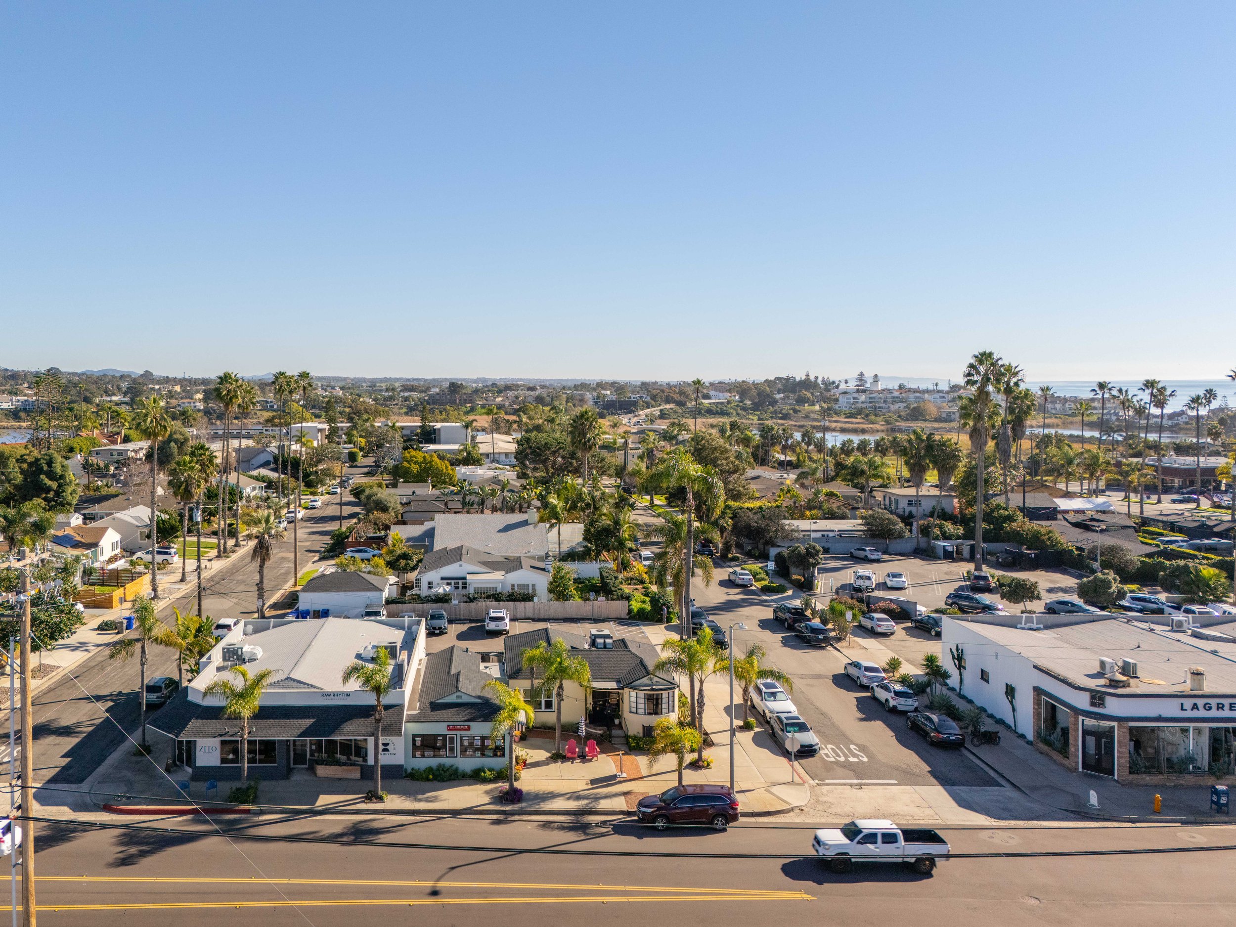 A panoramic view of a cityscape featuring residential houses, palm trees, streets, and parking lots under a clear blue sky.