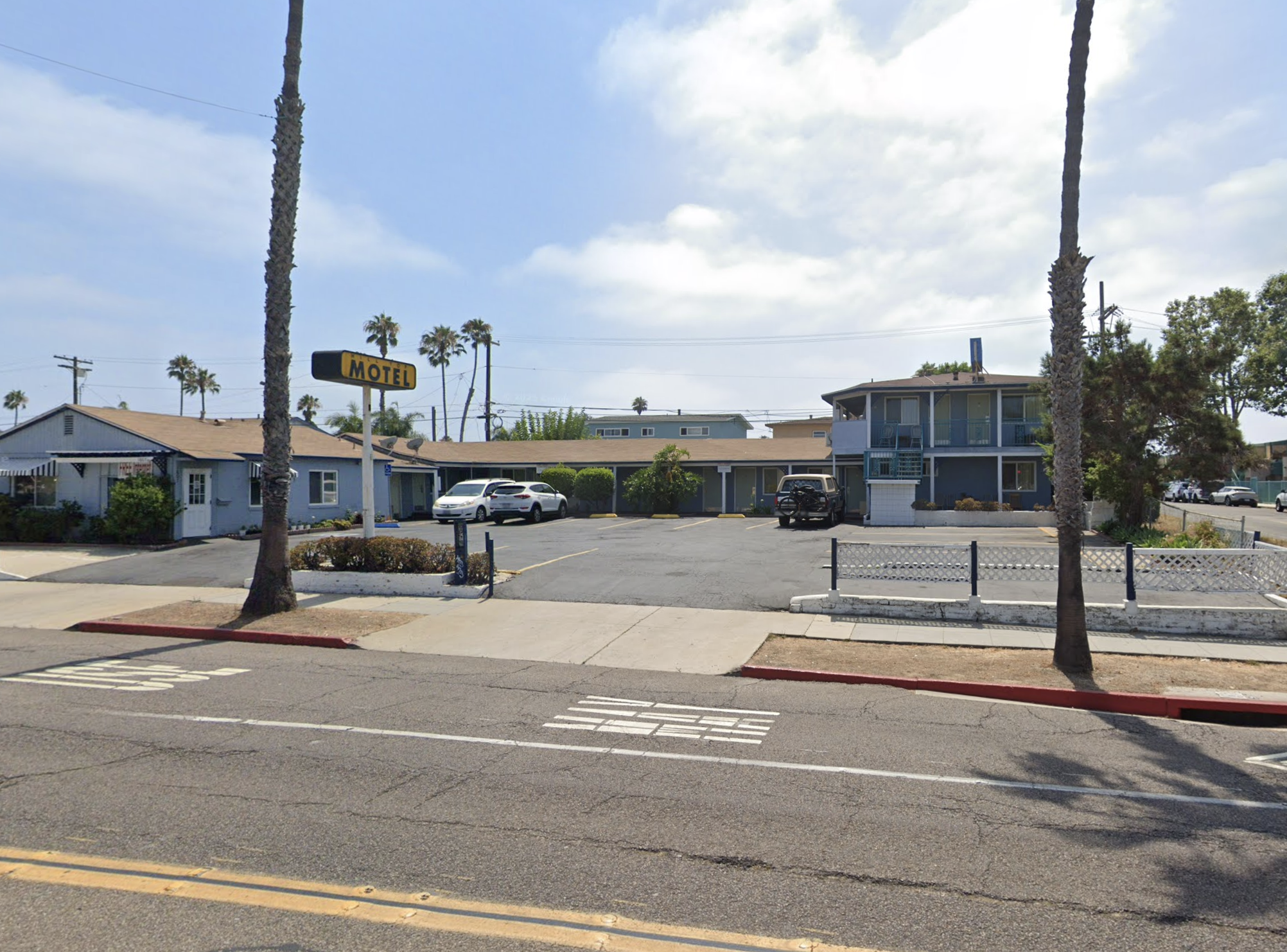 A parking lot with several cars parked and a motel in the background, two tall palm trees in the foreground, and smaller palm trees in the background under a partly cloudy sky.