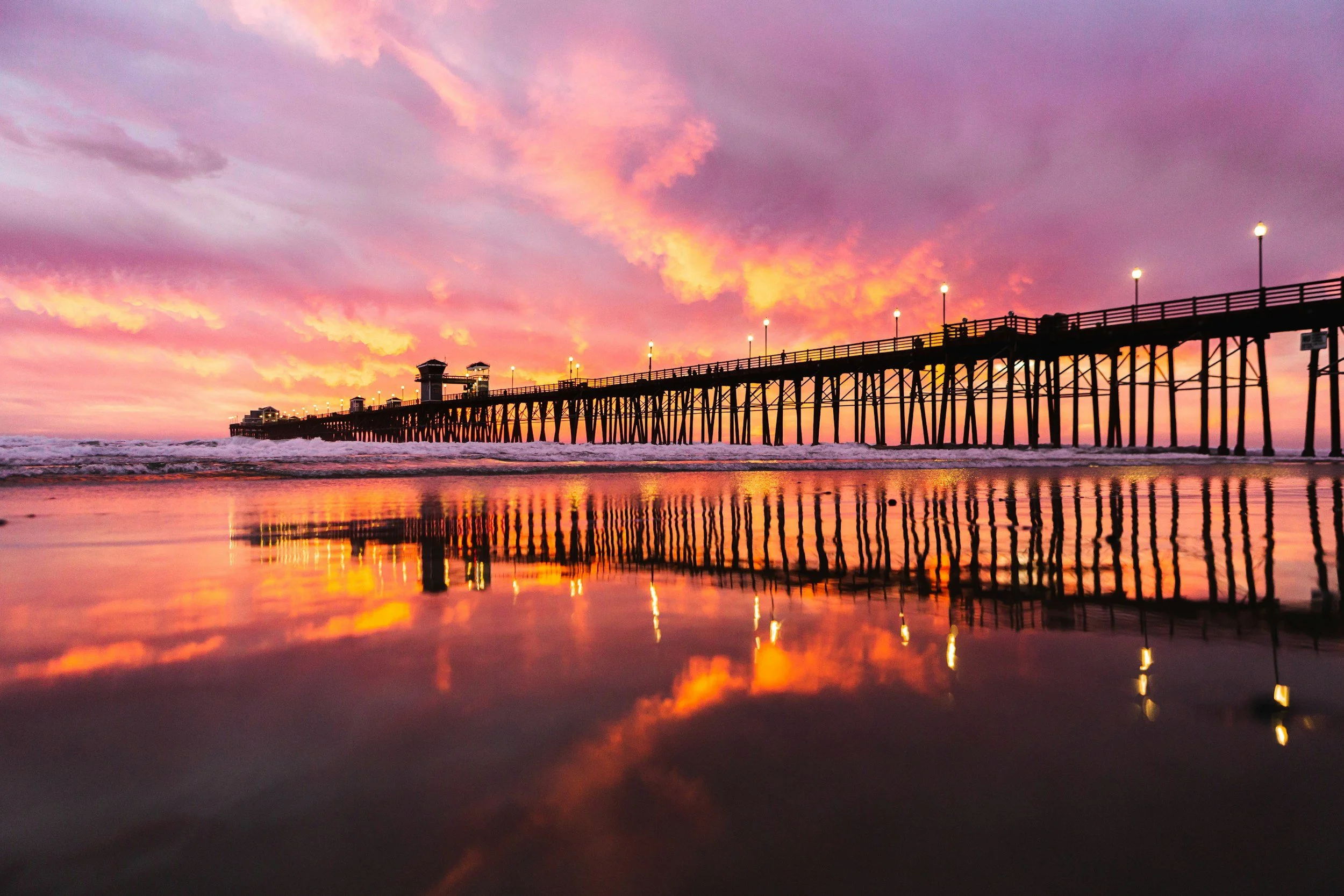 Oceanside pier extending over the ocean at sunset with colorful orange, pink, and purple clouds in the sky, and their reflection visible in the water below.
