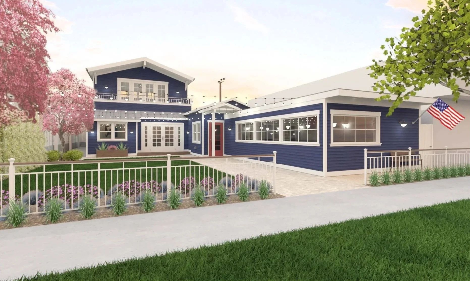A modern two-story house painted in navy blue with white trim, large windows, and a front porch. The yard has a white fence, green grass, flowering plants, and pink trees. An American flag is displayed on a pole near the house.