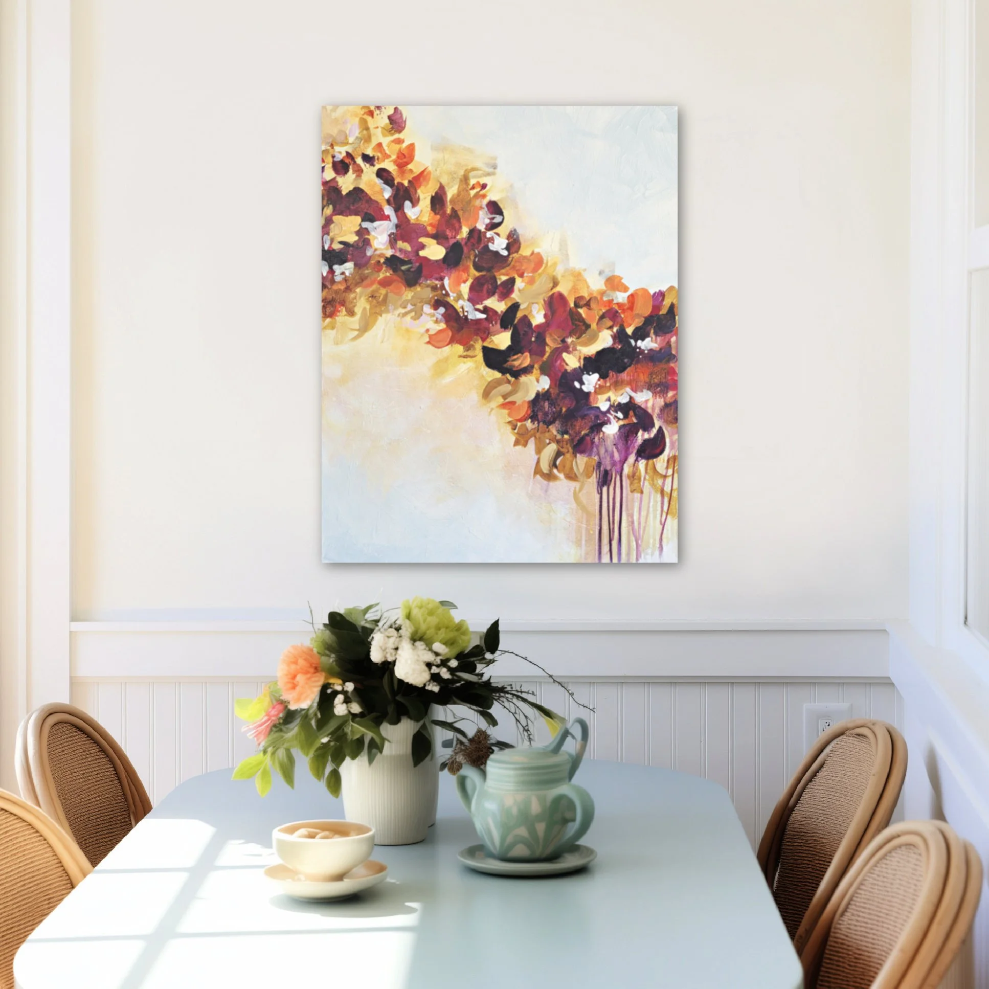 A dining room with a light blue table, featuring a centerpiece of white, green, orange, and yellow flowers in a white vase. There are three beige chairs with woven backrests around the table. In the background, there is a white wall with a large abstract painting with colorful brushstrokes. On the table, there are three small bowls and two green vases.