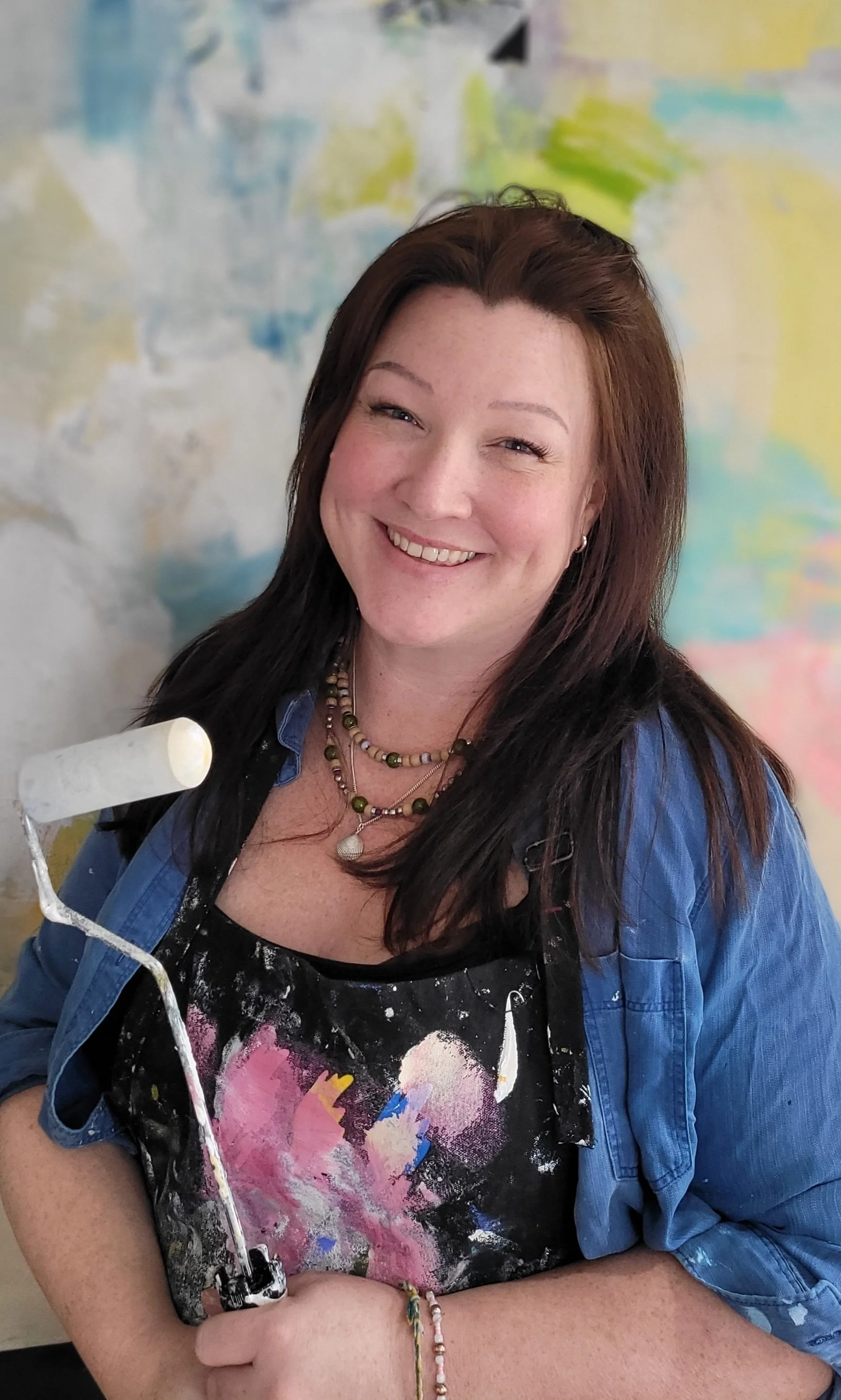 A smiling woman with dark brown hair and light skin holding a paint roller in front of an abstract, colorful wall.