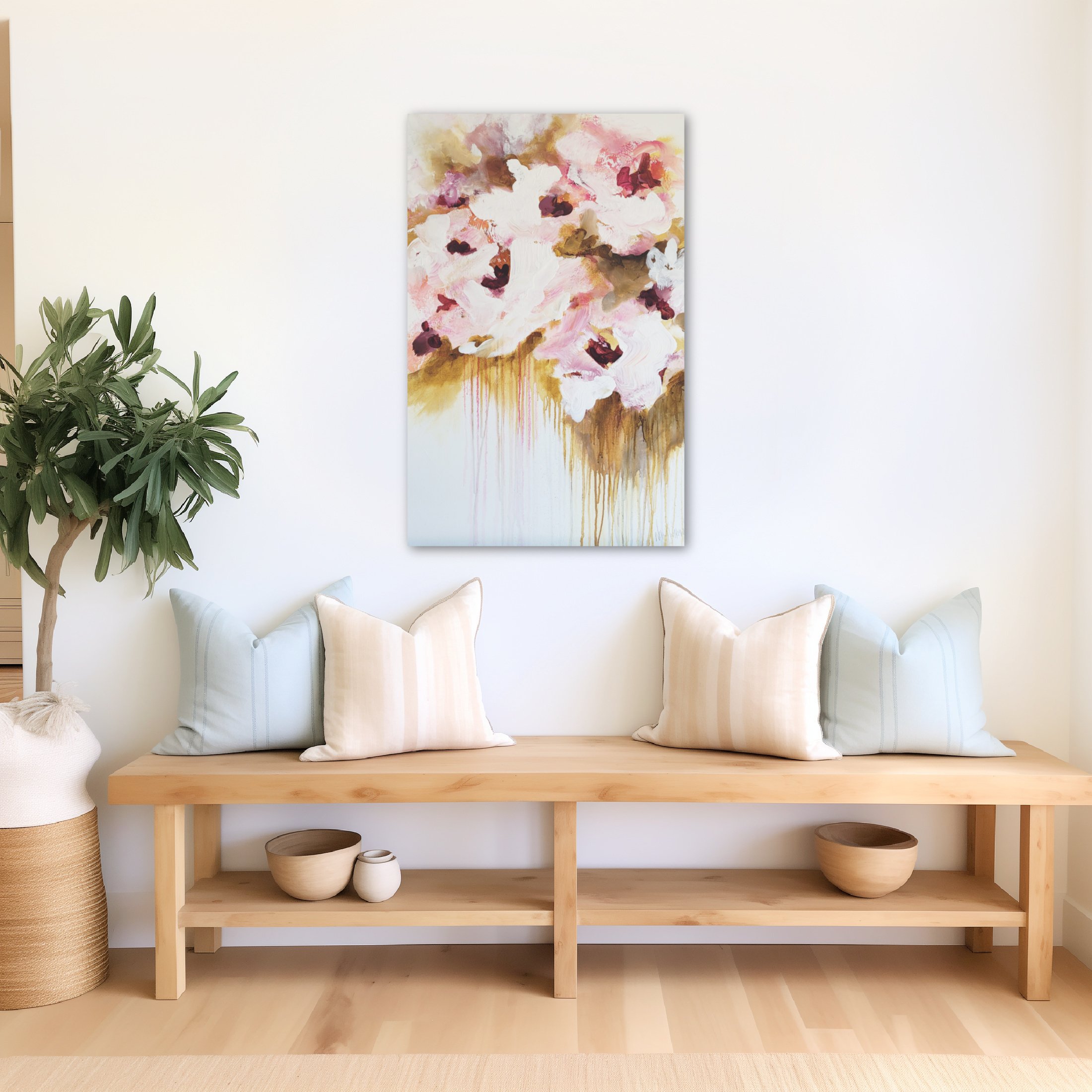 A simple wooden bench with a lower shelf, placed against a white wall. The shelf holds several white ceramic bowls and a small jar. There are light-colored cushions on the bench. To the left, a large green plant with broad leaves in a woven basket. Above the bench, a colorful abstract painting with pink, white, brown, and gold tones is hanging on the wall.