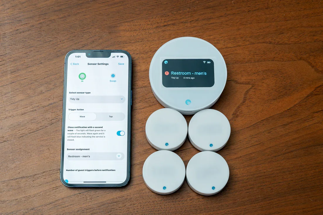 Smartphone displaying sensor settings next to several circular motion sensors and a digital display showing 'Restroom - men's' on a wooden surface.