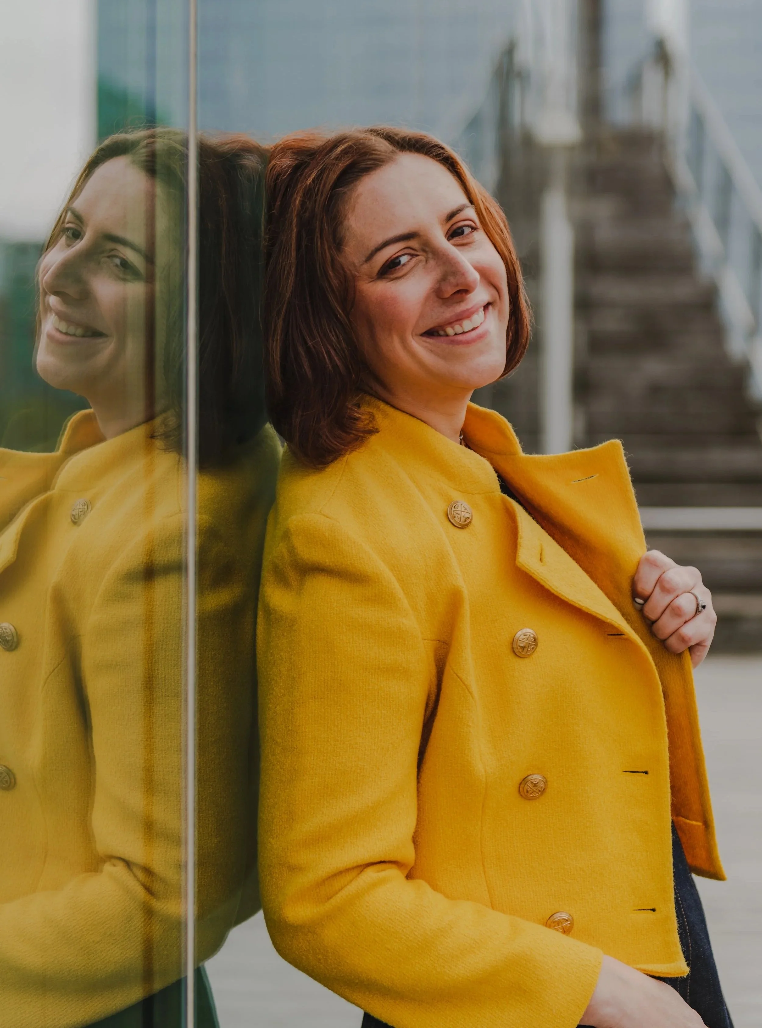 A woman with short brown hair wearing a yellow coat standing next to a glass wall, smiling, with her reflection visible in the glass.
