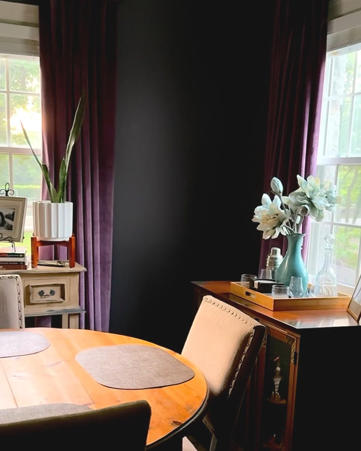 Moody, cozy, rustic modern dining room with plush purple velvet curtains, dark walls, neutral wooden dining table, vintage cabinet and glassware and other collected items.