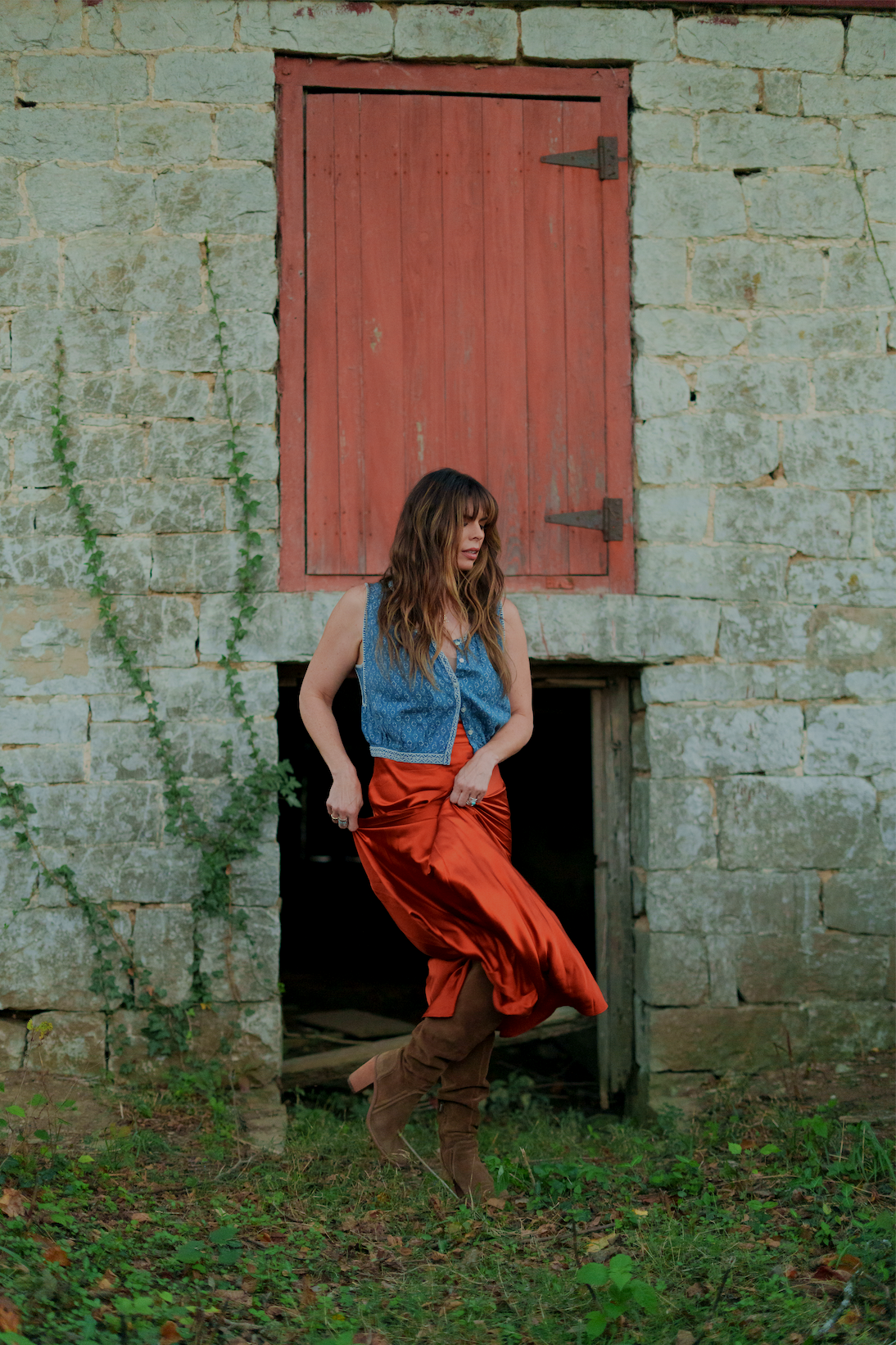 Woman with brown hair stomping through grass in brown boots, burnt orange silk skirt and blue tank top in front of rustic stone wall