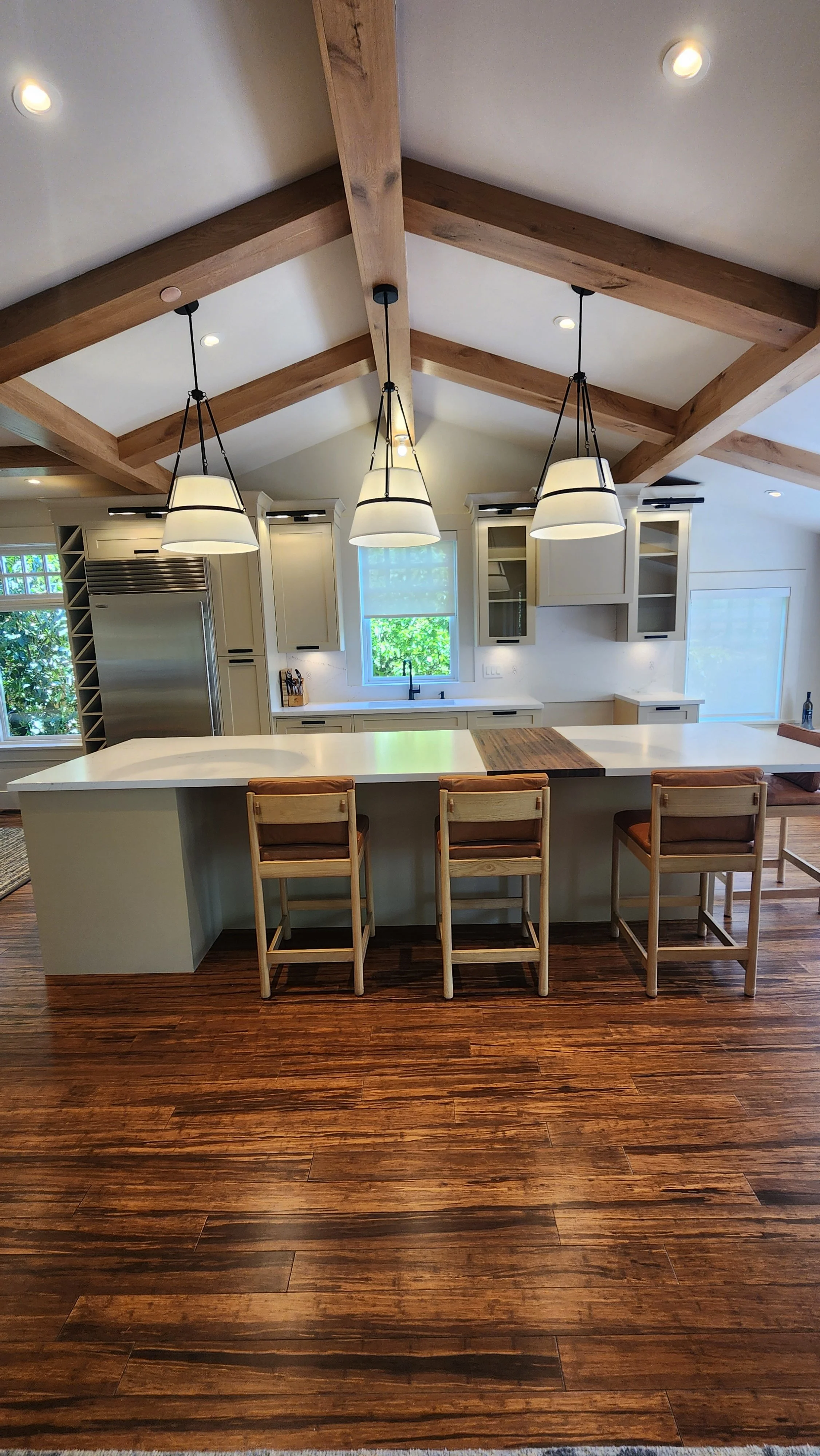 White kitchen cabinetry and island beneath exposed timber beams, with wood stools and pendant lights.