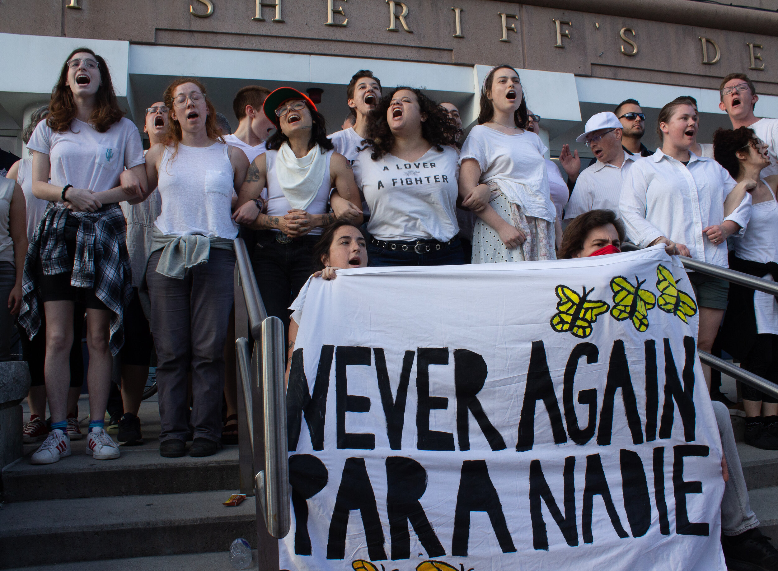 Group of people wearing white, with mouths open as if singing, holding a banner that reads "Never AGain Para Nadie". The building behind them says "Sheriffs Dept"