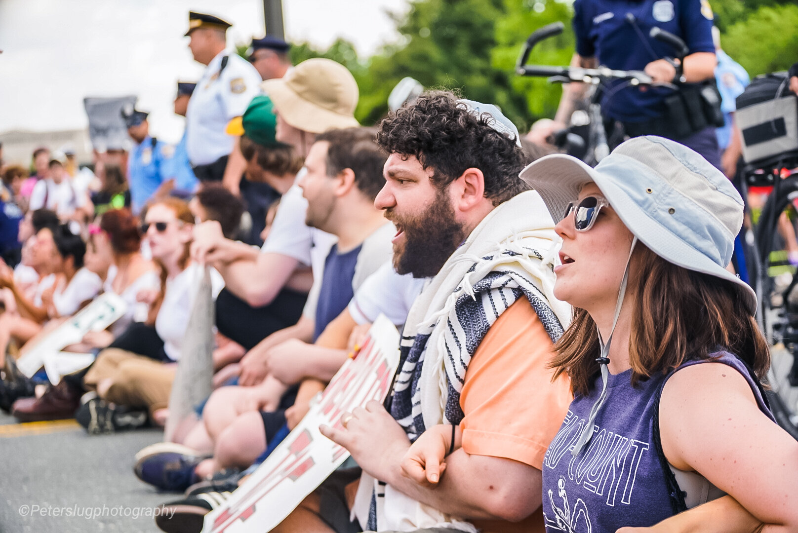 row of people sitting on the ground, one wearing a tallis (Jewish prayer shawl), with police standing in background