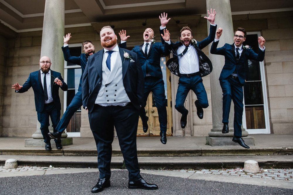 groom and groomsmen pose for jumping portrait in front of haigh hall steps