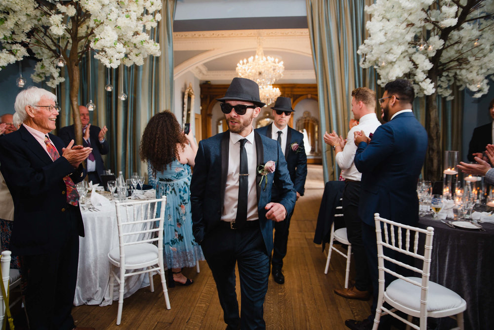 best men wearing hats and sunglasses being clapped into wedding breakfast room