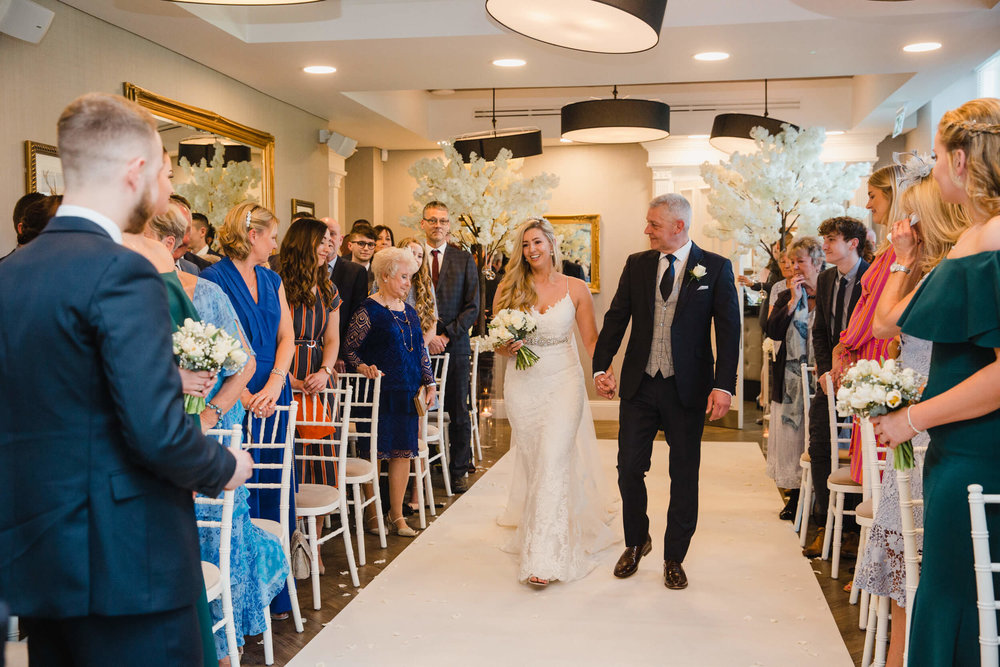bride and father walk down aisle together surrounded by wedding party