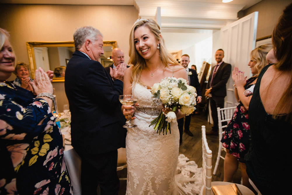 smiling bride holding champagne and bouquet entering wedding breakfast room