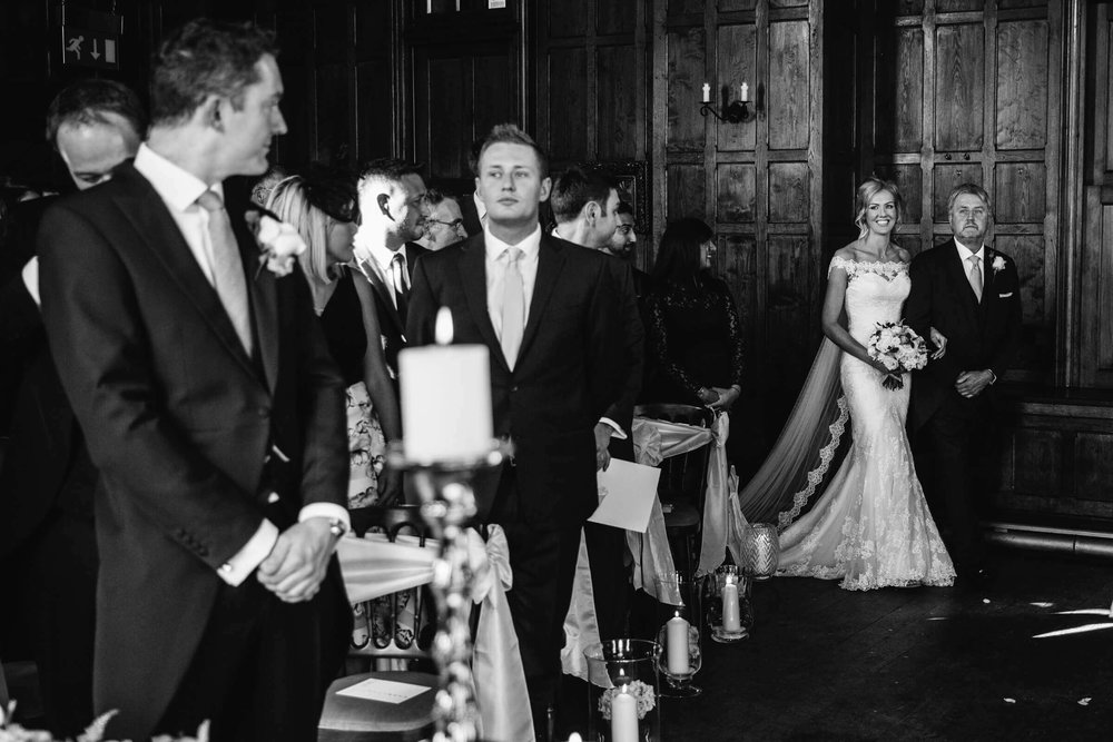 black and white photograph of bride entering ceremony room to processional with groom in foreground