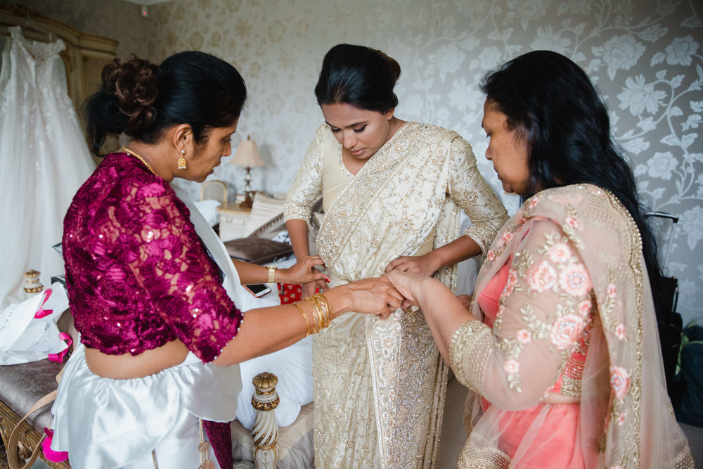 bride being dressed into wedding gown by bridesmaid and mother