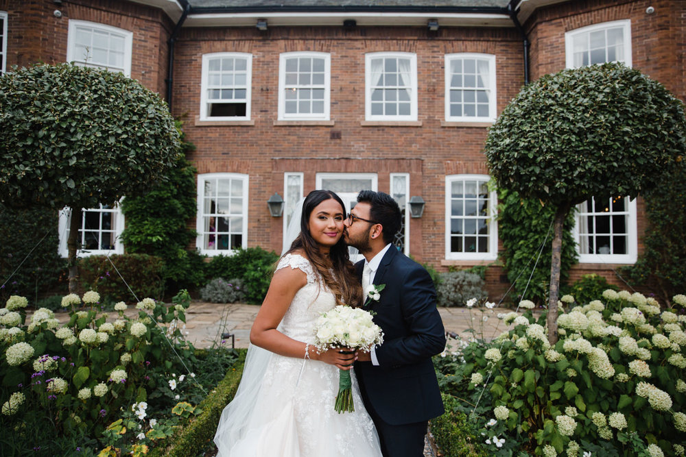 bride looks at camera while groom kisses her cheek with delamere manor estate in the background