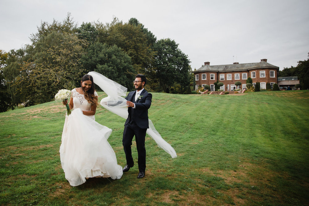 wind blows brides wedding dress veil as groom laughs to help her in the gardens