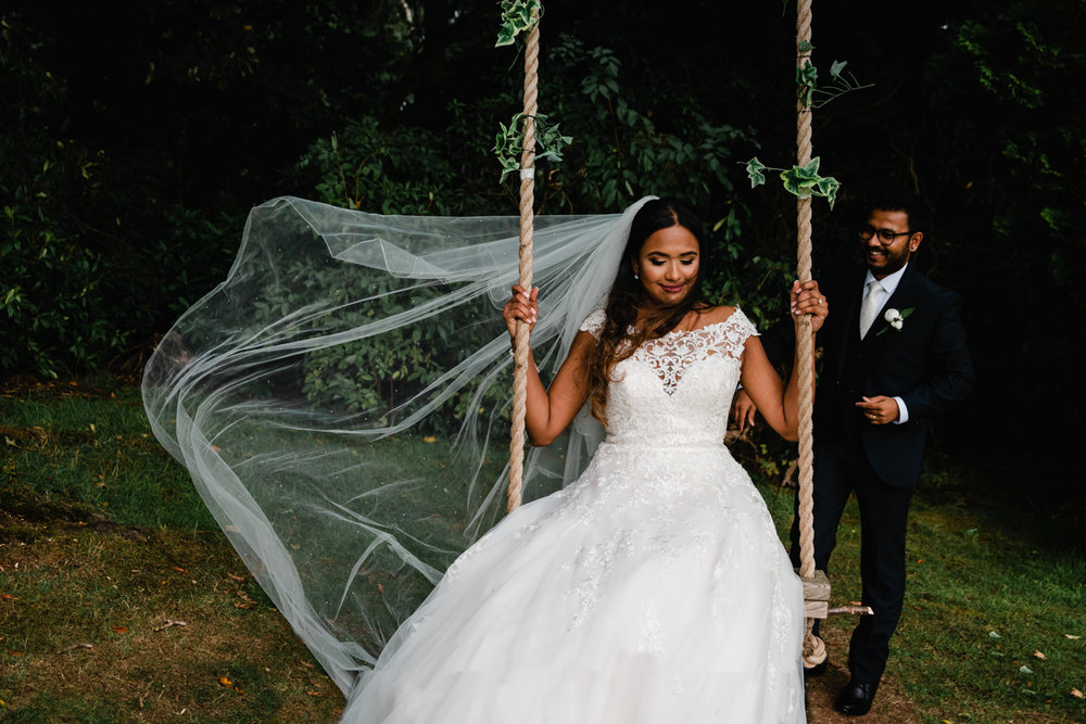 bride on swing as wedding dress veil blows in the air and groom looks on in the background