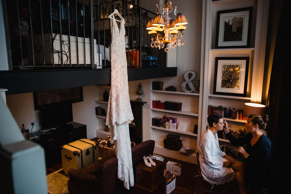 Copy of wedding dress hung from bannister while bride has make up applied in bridal suite at great john street hotel