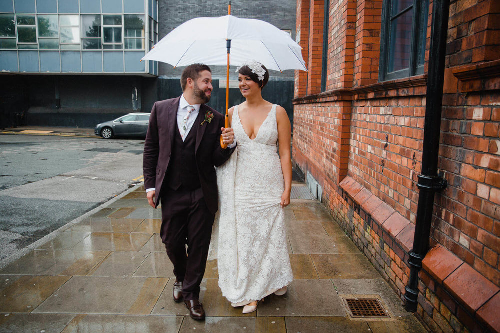 Copy of newlyweds walk down pavement for posed portrait next to alley towards camera holding umbrella 