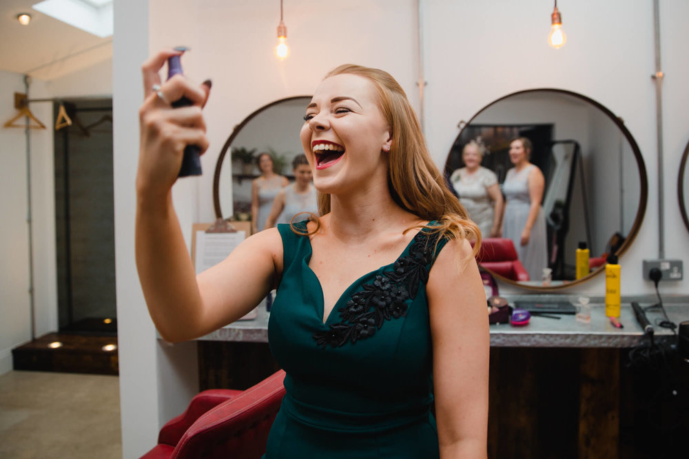 natural funny moment with bridesmaid spraying face as bridal party look on in the mirror