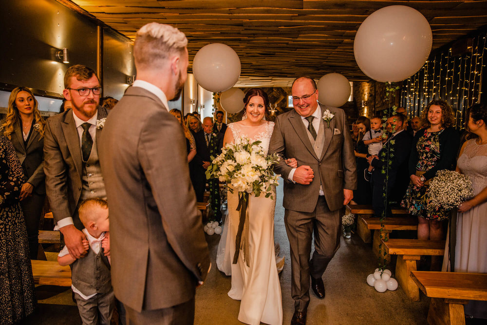 father of bride walks daughter down aisle to wedding processional as groom looks on in foreground